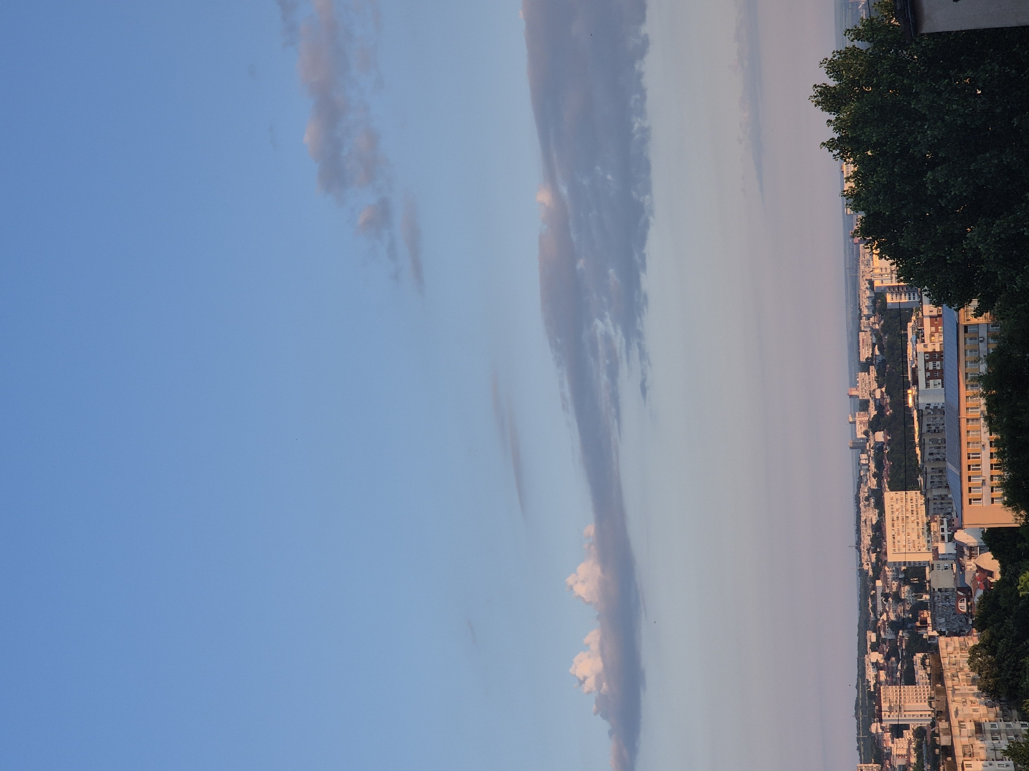 An aerial view of an urban landscape under a blue sky with scattered clouds. The city appears to be densely populated with numerous buildings of varying heights and architectural styles. Some buildings are modern high-rises, while others are older, lower-rise structures. Green trees are interspersed throughout the city, particularly in the foreground and along the edges of buildings, suggesting a mix of urban development and natural elements. The lighting indicates it could be late afternoon or early evening, with the sky exhibiting soft hues of pink and orange near the horizon. The overall scene depicts a typical cityscape with a serene sky above.