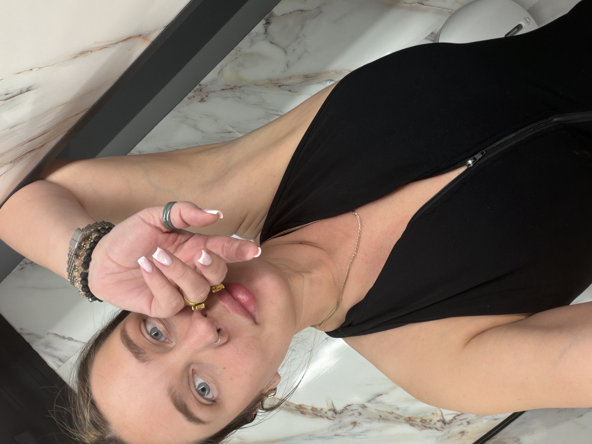 The image is a low-angle perspective selfie of a Caucasian woman with fair skin, light blue eyes, and light brown hair. She is wearing a black top with a zipper that is partially unzipped, a gold chain necklace, a beaded bracelet on her left wrist, multiple rings on her fingers, and hoop earrings. The woman's left hand is raised, partially obscuring her face. She has manicured nails with white tips. Her lips are full and appear to be slightly glossy. She has a nose ring on her left nostril. The background appears to be a surface with a marble pattern. There is a dark gray or black strip along the left side of the image, possibly a wall or edge of furniture. There is also a white, rounded object in the top right corner of the image, which is possibly a piece of equipment. The setting appears to be indoors. There is no evidence to suggest that the image was taken in Moscow, Russia.