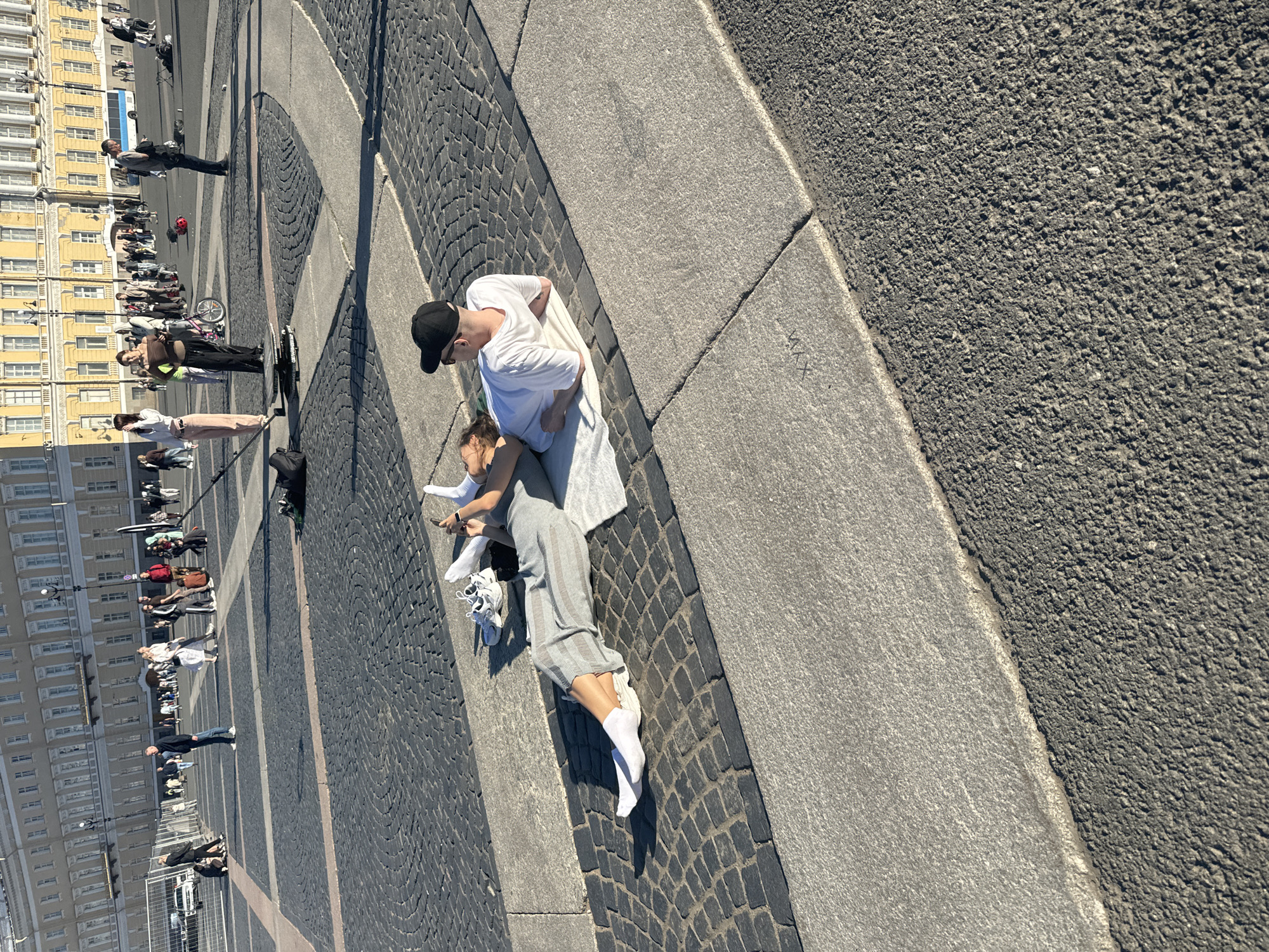 The image depicts a couple lying on a white blanket on a paved public square in Saint Petersburg, Russia, possibly Palace Square, given the architectural style of the building in the background. The woman, wearing a grey jumpsuit and white socks, is looking at her phone, while the man, wearing a white t-shirt and a black baseball cap, rests his head on her shoulder. White sneakers are placed beside them. The square is paved with dark cobblestones and large grey stone slabs, and in the background, a large, light-colored building with numerous windows and classical architecture dominates the scene, characteristic of many St. Petersburg landmarks. Other people are visible in the distance, walking or standing in the square. The lighting suggests it is daytime, with strong sunlight casting shadows.