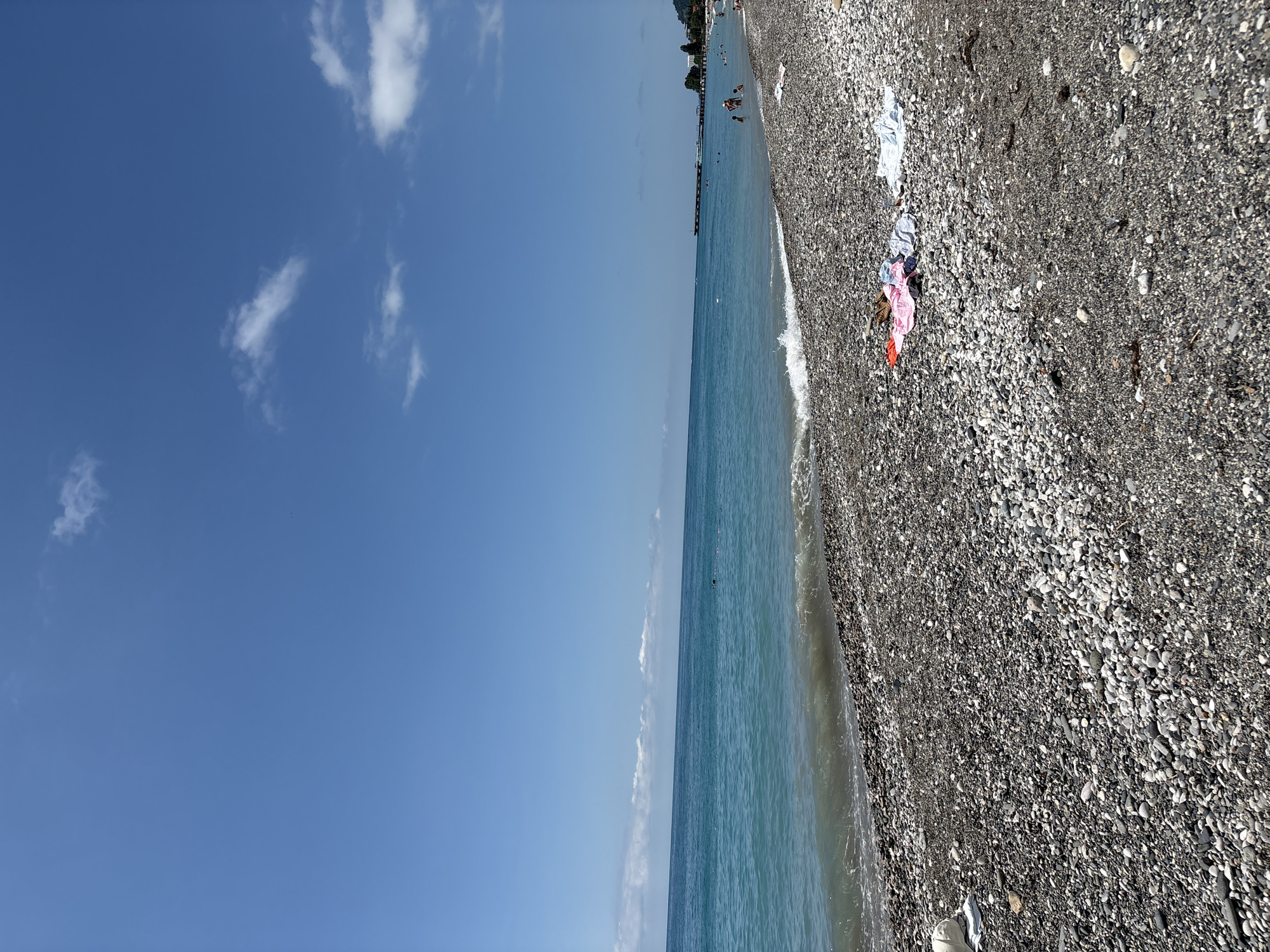 The image shows a sunny day at a pebbled beach in Abaata, Abkhazia. The sea is a vibrant turquoise, with gentle waves lapping the shore. The sky is a clear, bright blue, with a few wispy clouds scattered across it. In the foreground, the beach is covered in smooth, grey and white pebbles. Several pieces of clothing, including a pink towel, a brown bag, a blue and white striped shirt, and a white towel, are laid out on the pebbles. In the distance, people can be seen swimming and relaxing in the sea, and a long pier extends out into the water. The shoreline curves into the distance, with trees and buildings visible on the hillsides.