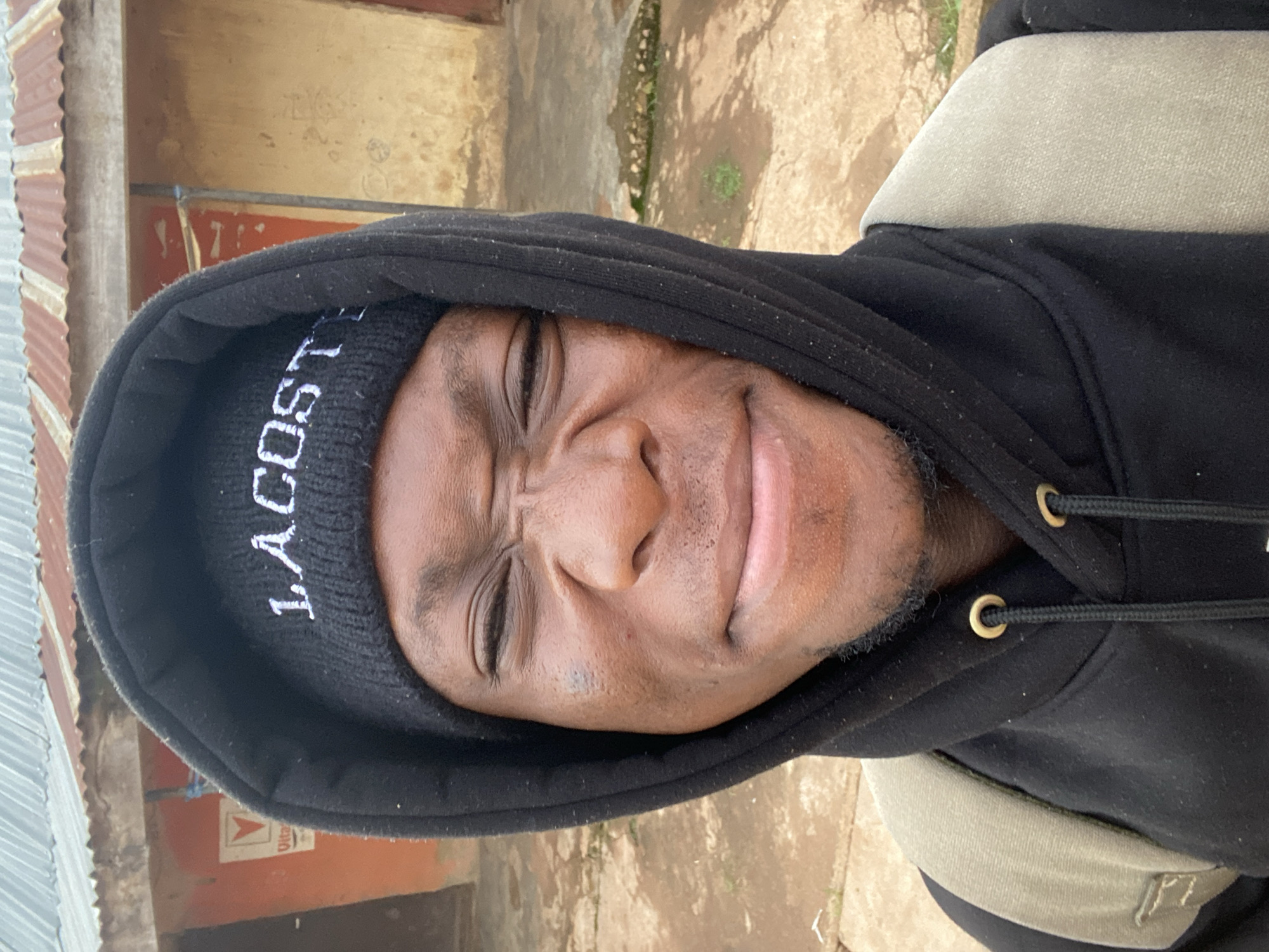 A young Black man with his eyes scrunched shut and a grimace on his face is shown in a close-up, slightly high-angle selfie. He is wearing a black hoodie with the hood pulled up and a black beanie that says "LACOSTE" in white lettering. A tan backpack strap is visible over his left shoulder. The background appears to be an outdoor setting with a corrugated metal roof and a building with an orange wall and a tan wall. The ground is dirt and appears wet in places. The overall impression is that this image was taken in Vom, Nigeria, possibly in a residential or commercial area.