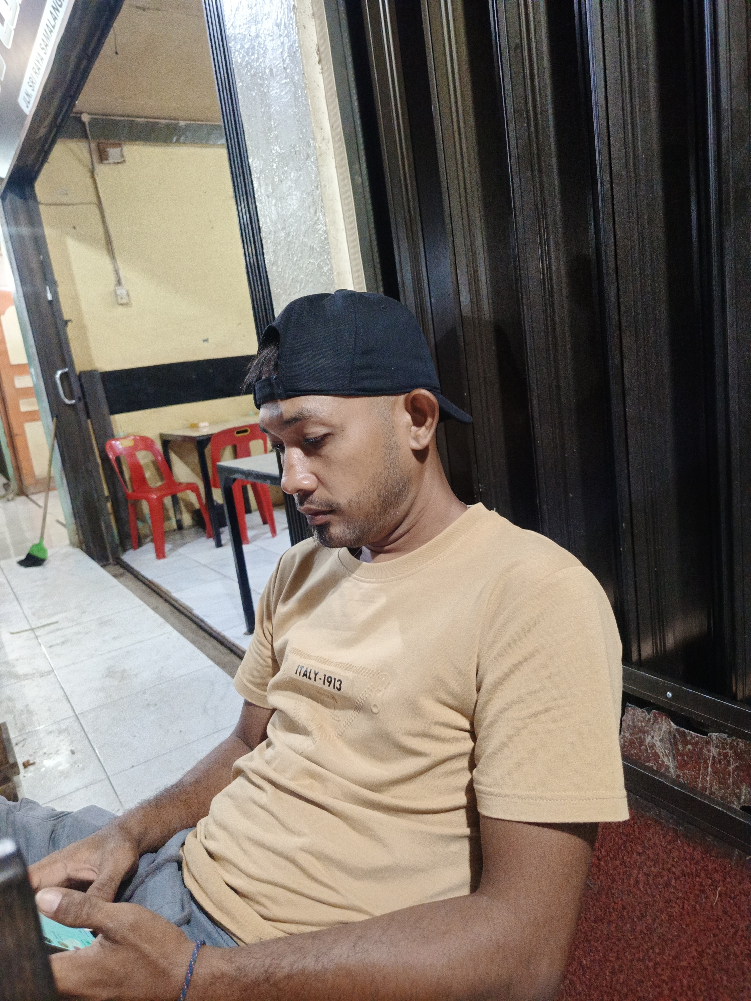 An adult male, wearing a black baseball cap, a light brown short-sleeved t-shirt with "ITALY-1913" text, and grey drawstring pants, is seated and looking downwards at a smartphone held in his hands. A blue beaded bracelet is visible on his left wrist. The scene takes place in Sangso, Indonesia. The background to the man's right features a dark, vertically corrugated metal surface. To his left, through an open doorway, an interior space with light yellow walls is visible, furnished with two red plastic chairs and two tables with dark tops. The floor consists of light grey square tiles. A brown broom with a green base leans against the wall. Partial signage above the doorway reads "JLN SRI RAYA SAMALANG".