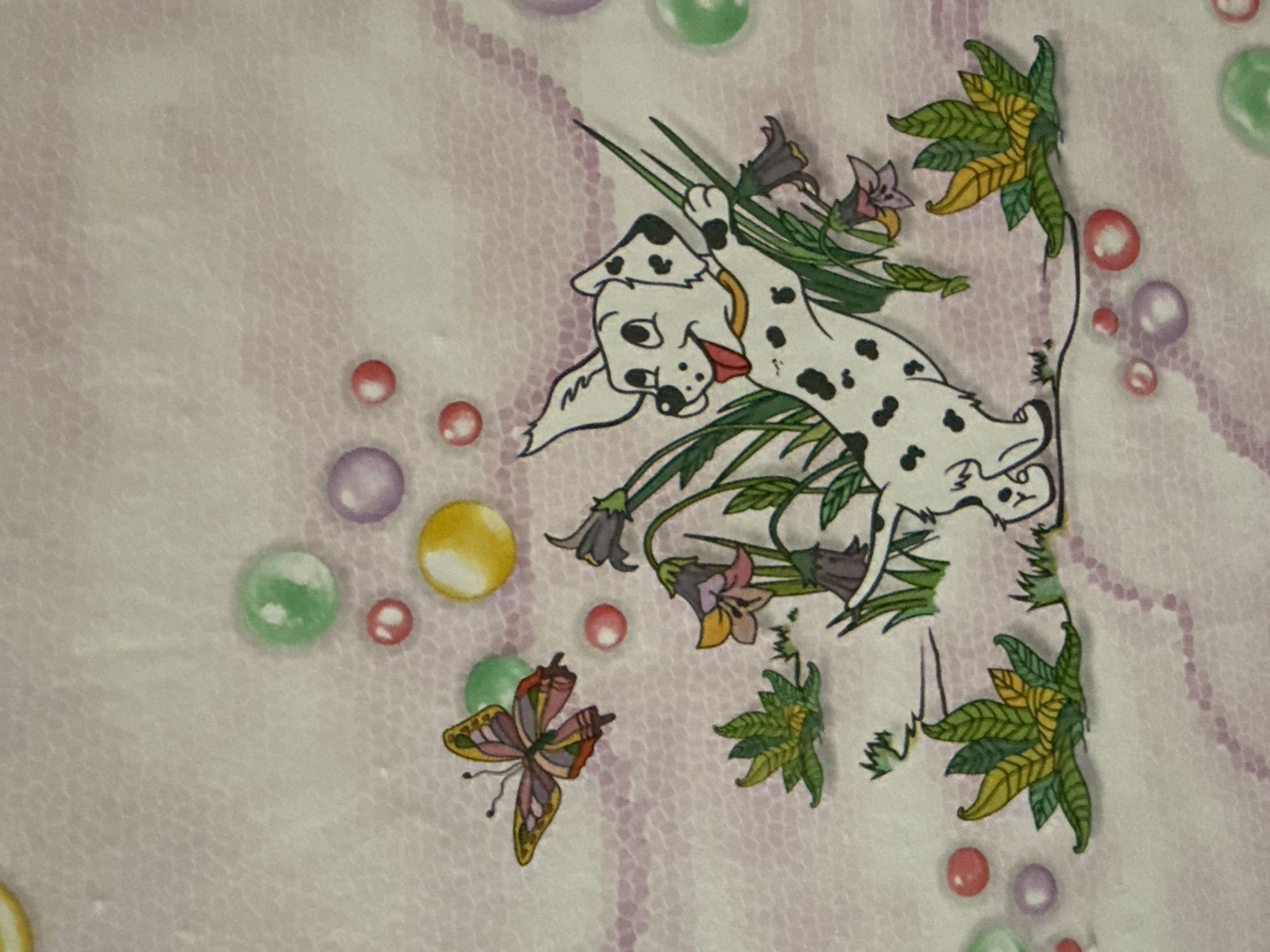 The image is a close-up, overhead shot of a piece of fabric with a cartoon illustration. The illustration features a black and white spotted dog with its tongue out, reaching up a stalk of grass. The dog is surrounded by stylized green foliage and purple flowers with yellow centers. To the left of the dog, a colorful butterfly with orange, pink, and green wings is depicted in flight. Scattered around the illustration are several large, translucent colored circles in shades of green, purple, and yellow, resembling bubbles. The background of the fabric has a faint textured pattern in pink and white. There are no identifiable landmarks or elements that would suggest a specific location within the city of Bălți, Moldova. The scene is entirely fictional and illustrative.