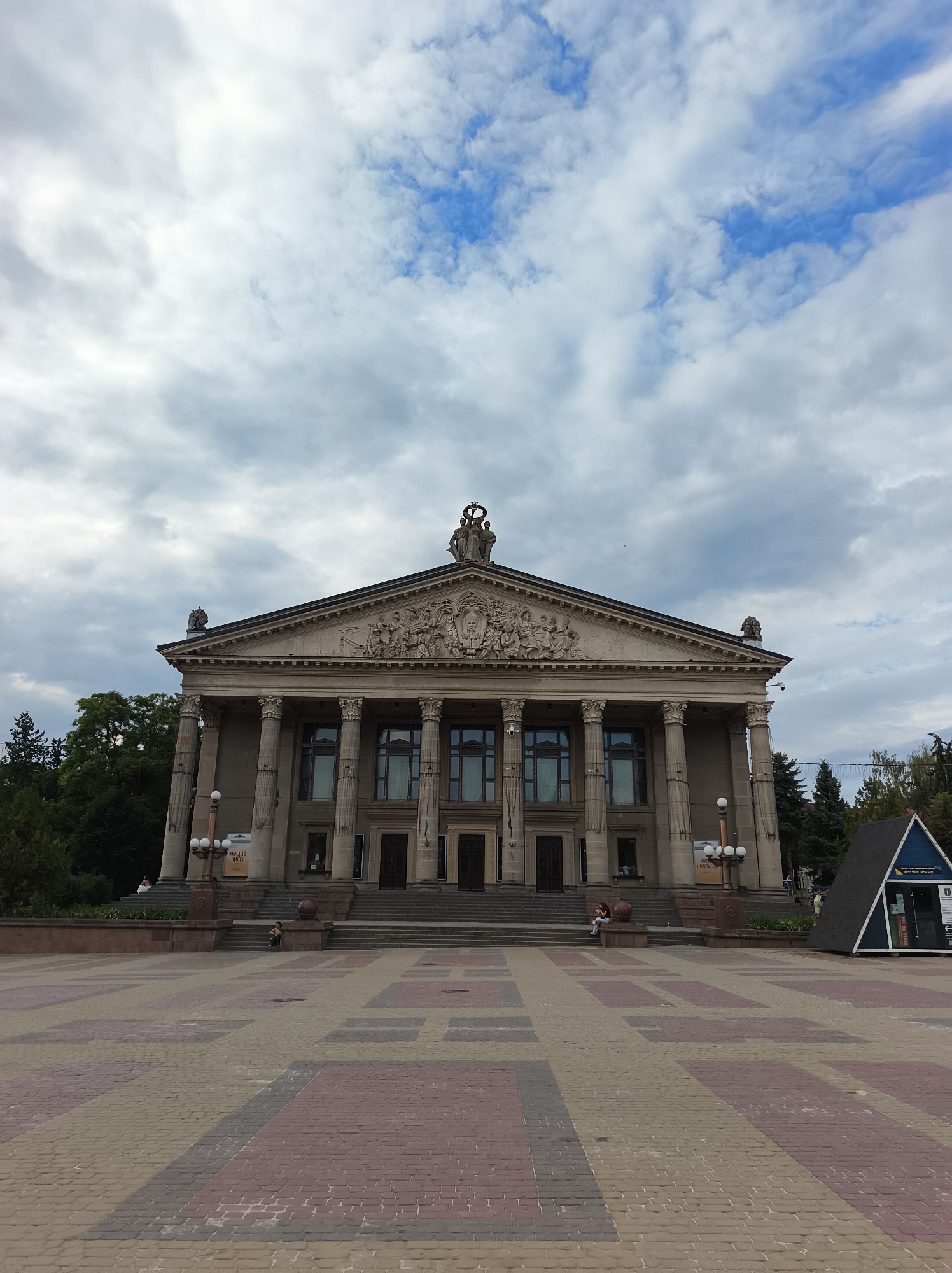 The image shows the Ternopil Regional Philharmonic, a grand neoclassical building located in Ternopil, Ukraine. The building features a prominent portico with Corinthian columns, a pediment adorned with sculptures, and ornate friezes. A wide, paved plaza stretches out in front of the philharmonic, with a patterned arrangement of tan, red, and gray paving stones. Several people are visible in the scene, including a few individuals sitting on the steps of the building and one person walking in the plaza. The sky above is partially cloudy, with patches of blue peeking through. To the right of the philharmonic, a small, triangular-shaped structure with signage is situated in the plaza. The overall impression is one of a public square in front of a significant cultural institution, with a mix of architectural grandeur and everyday human activity.