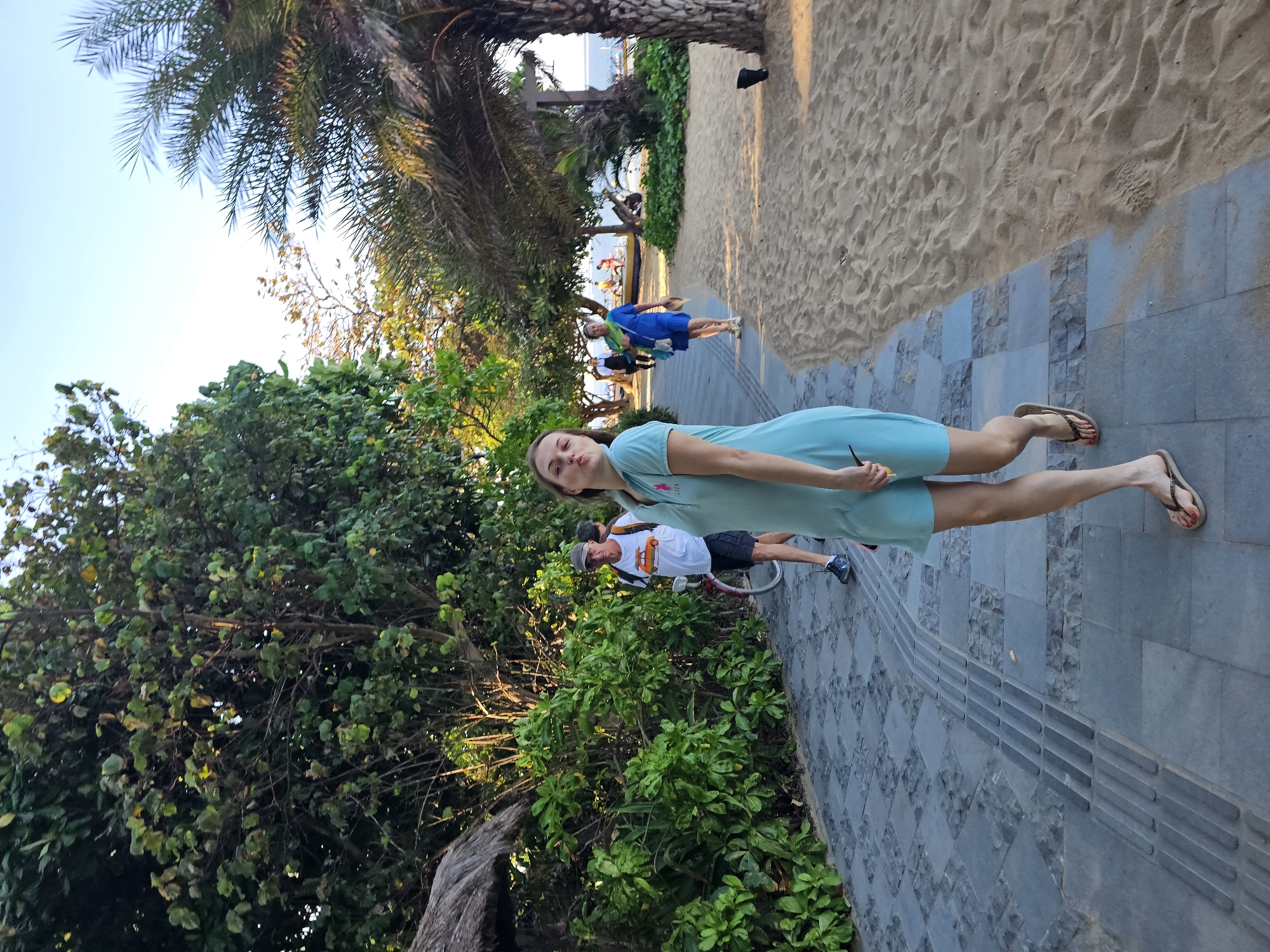 A group of people are walking along a paved path next to a sandy beach. The path is made of grey stone tiles and has tactile paving. In the foreground, a young woman in a light blue dress is making a kissing face at the camera. Behind her, two men are walking; one is wearing a white t-shirt with a graphic and black shorts, and the other is wearing a dark t-shirt and black shorts. Further down the path, a woman in a blue dress and a scarf is walking away from the camera. Palm trees and lush green foliage line the path, and in the distance, the ocean is visible under a clear blue sky. This scene is likely at Kuta Beach in Denpasar, Indonesia, a popular tourist destination known for its beachfront walkways and palm-lined shores.