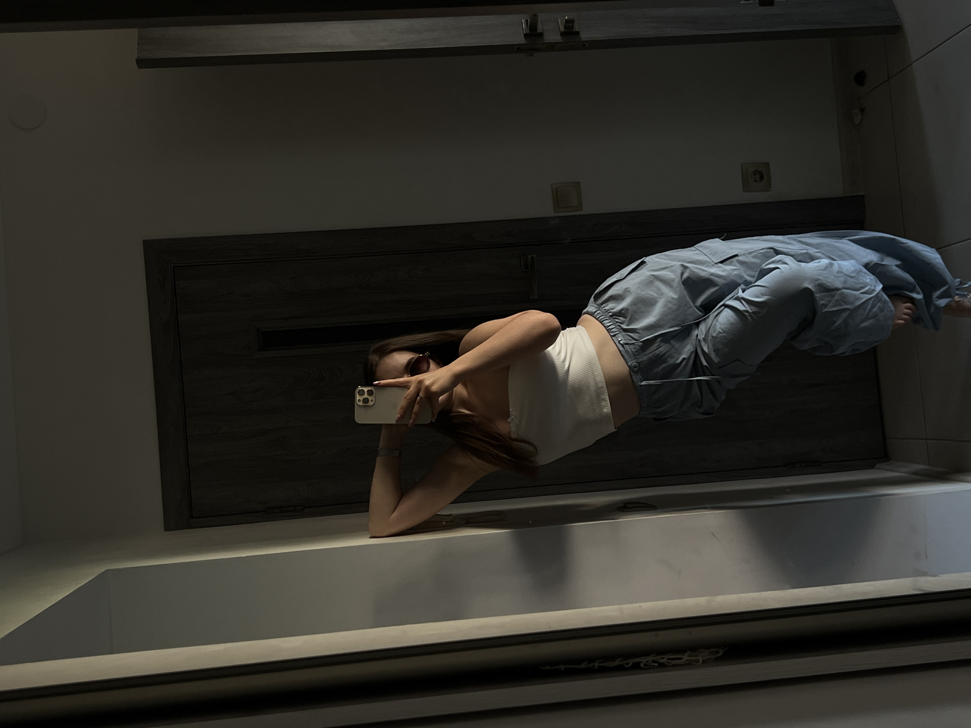 A woman is captured in a mirror selfie, lying on her back and holding a smartphone to take the photo. She is wearing a white crop top and light blue cargo pants. Her sunglasses are on her head. The background appears to be an indoor setting, possibly a bathroom or a room with a large mirror, given the reflective surface dominating the lower half of the image. The room is dimly lit, suggesting it might be evening or night. The wall behind her features a dark wooden door and a light-colored outlet. The overall mood is casual and relaxed. There is no visible text in the image.