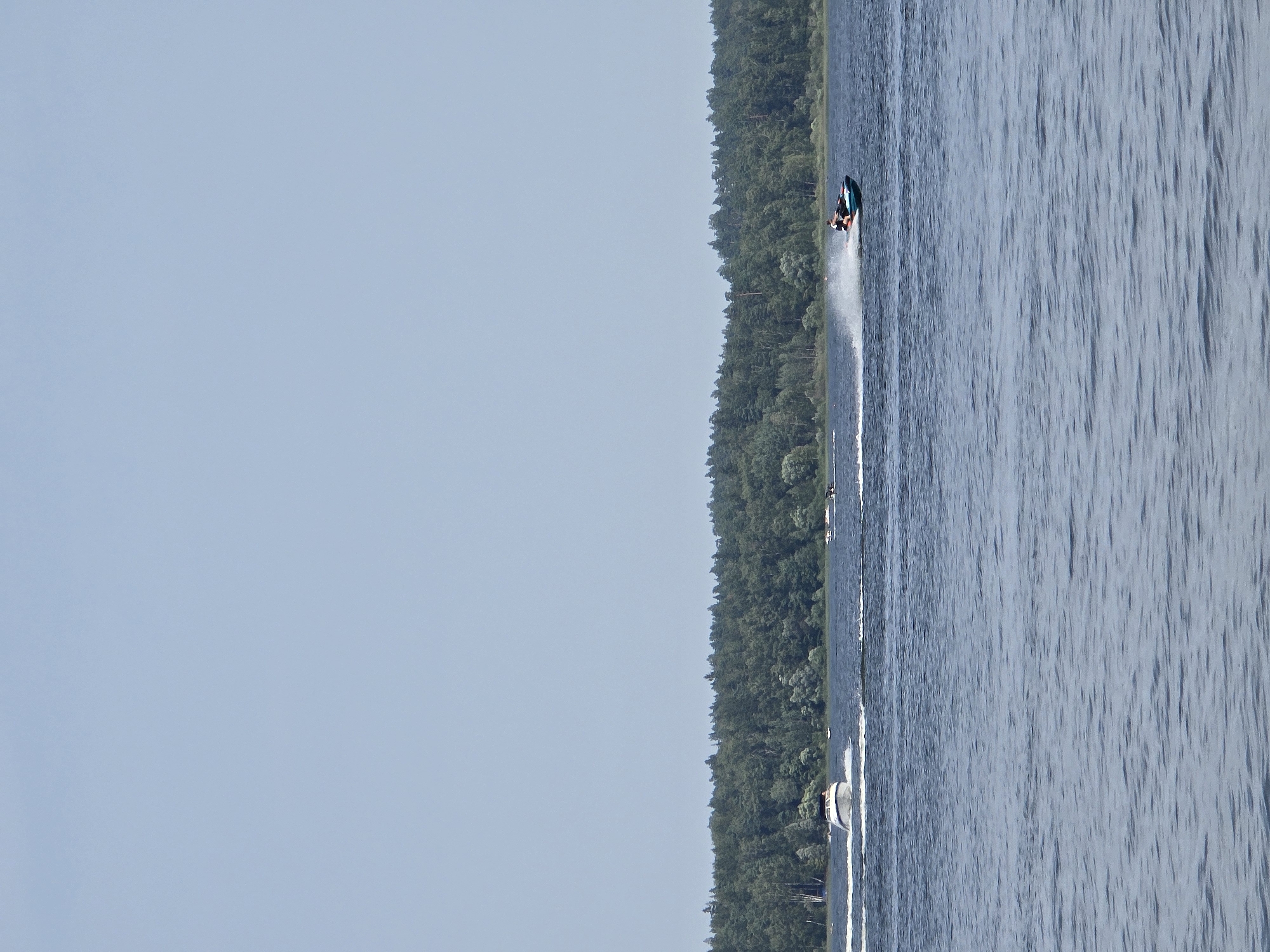 The image depicts a wide body of water, likely a lake or river, with a dense, green forest lining the distant shore. The sky is a pale blue, largely cloudless. In the foreground, the water is rippled. Two watercraft are visible on the water. On the left, a white motorboat is speeding across the water, leaving a white wake. On the right, a person is riding a turquoise jet ski, also creating a significant spray of water. The overall scene suggests a recreational activity taking place on a clear day. The location is identified as посёлок гидроузла имени Куйбышева, Russia.