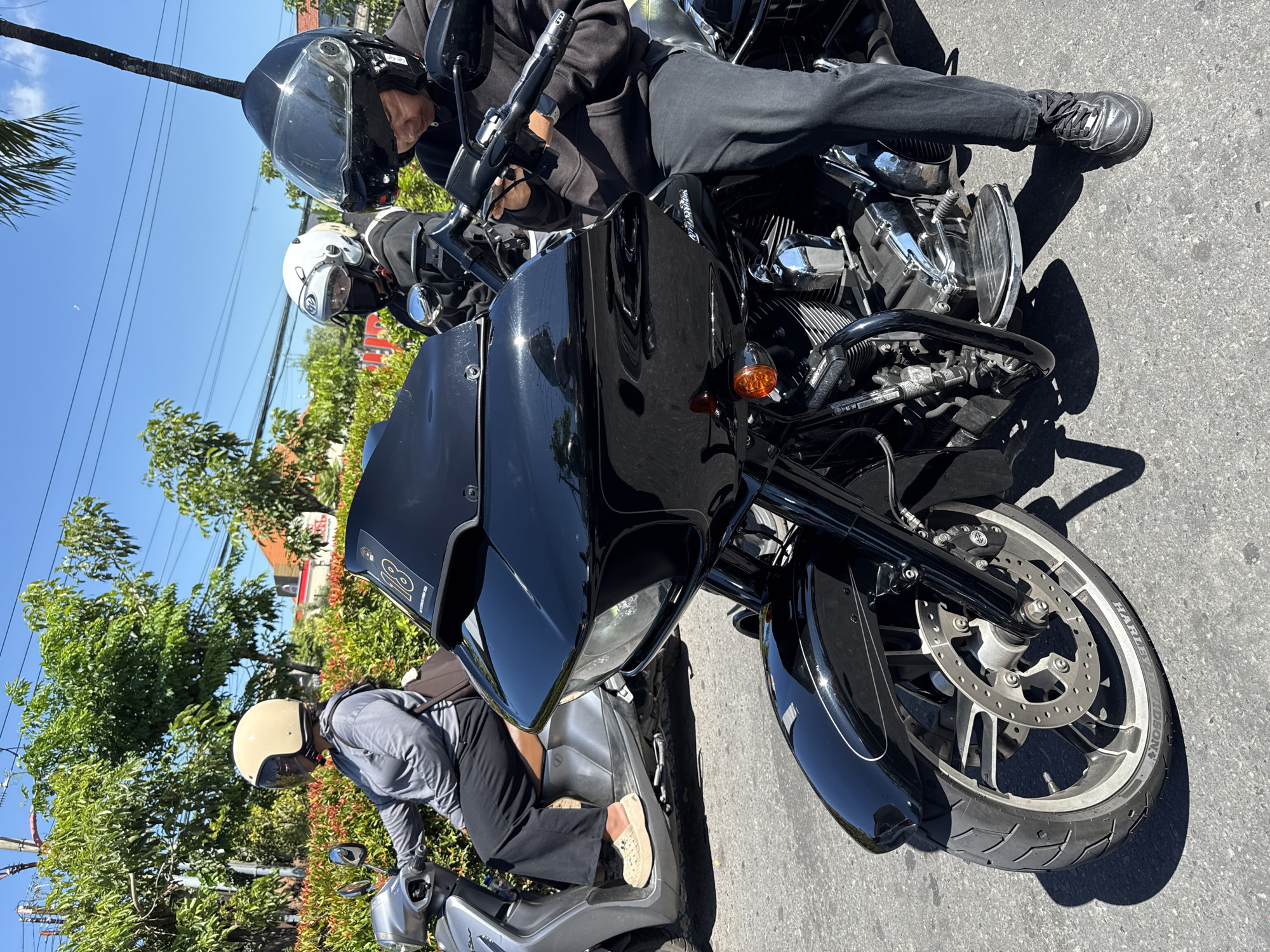 The image depicts a street scene in Denpasar, Indonesia, showcasing a black Harley Davidson motorcycle positioned prominently in the foreground. The motorcycle is flanked by other motorcycles, including a smaller silver scooter, which is being ridden by a person wearing a beige helmet, a gray shirt, and dark pants. The person on the Harley Davidson wears a full black helmet and dark clothing, sitting astride the bike. A second motorcyclist, clad in a white helmet, is situated behind the Harley Davidson rider. Lush greenery lines the street, with trees and bushes forming a backdrop. A blue sky and electrical wires are visible overhead. In the background, a partially visible building suggests the location is in an urbanized area. The motorcycles are parked on the paved asphalt road.