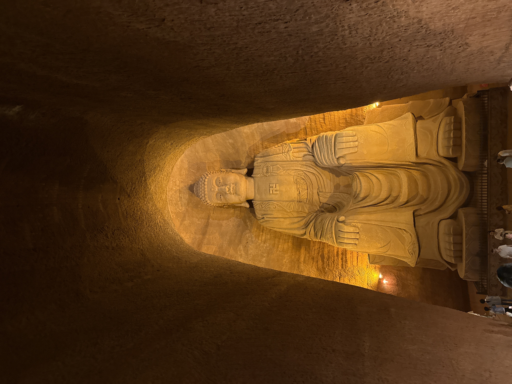 A massive, horizontally-oriented reclining Buddha statue, carved from light brown or sandy-colored stone, dominates the central and right portions of the image. The Buddha's head is positioned on the left, with its body extending to the right, depicting a serene and contemplative pose. A distinctive swastika symbol (Manji), representing peace and good fortune in Buddhism, is clearly visible on the Buddha's chest.
The statue is situated within a cavernous, dimly lit grotto or cave, carved into rough, dark brown and reddish-brown rock walls that curve above and to the left. Warm, golden-yellow artificial light illuminates the Buddha and select parts of the cave, casting dramatic shadows and highlighting the intricate carvings.
In the lower right corner, several small figures of people are visible, appearing to be visitors or tourists, observing the colossal statue from behind a railing. Their presence underscores the immense scale of the Buddha. The scene captures a moment within a significant spiritual or historical site, likely at night or in an interior space, emphasizing the grandeur and ancient craftsmanship of the carving. No other legible text is visible in the image.