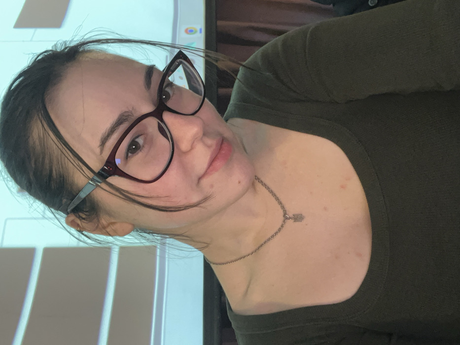 A young adult female is depicted in a close-up, chest-up shot, looking directly at the camera with a slight smile. She has dark hair, styled with some strands framing her face, and wears dark-rimmed glasses. A small gold-colored hoop earring is visible on her left ear. She is wearing a dark olive green long-sleeved top with a scoop neckline. A silver-colored chain necklace with a small pendant, resembling a trident emblem, is visible on her chest. Minor reddish blemishes are present on her upper chest and neck. The background is out of focus, revealing an indoor environment with a large, light blue illuminated screen, possibly a projector screen or display, showing white graphic elements and a cluster of colorful icons in the lower right. There is no visual information within the image to identify the specific location as Rafailovići, Montenegro.