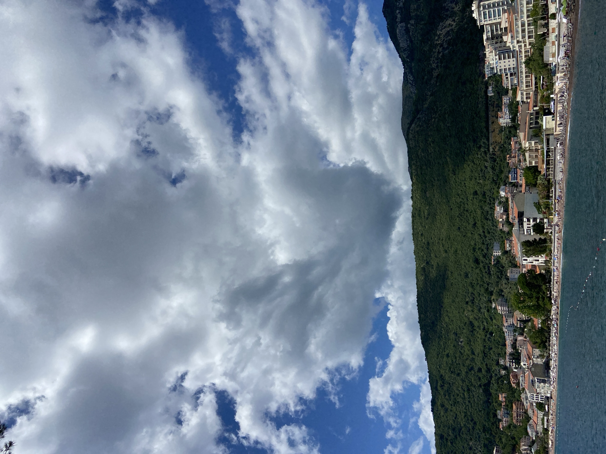 The image depicts a coastal scene in Petrovac centar, Montenegro. In the foreground, the dark blue water of the Adriatic Sea stretches across the bottom of the frame, with a thin rope barrier visible just below the surface. A gently sloping beach lined with numerous beach umbrellas and lounge chairs occupies the middle ground. Behind the beach, a dense cluster of buildings, consisting of hotels and residential structures with terracotta roofs, extends along the coastline. The buildings are nestled at the base of a steep, verdant mountain covered in thick green foliage. The upper portion of the image is dominated by a dynamic sky filled with a mix of white and dark gray cumulus clouds against a vibrant blue backdrop. The overall impression is a sunny day with potential for changing weather.