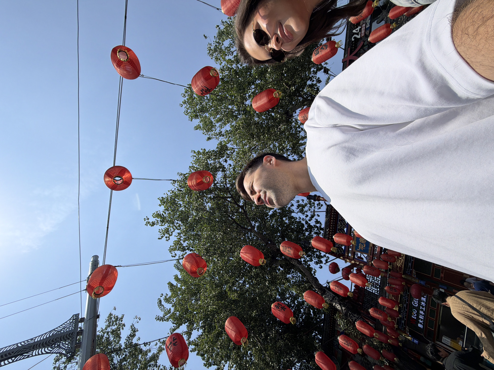 The image shows a low-angle view of two people in Dongcheng District, China. The man wears a white t-shirt and stands below a woman with sunglasses. Red Chinese lanterns hang from wires strung across the street, interspersed with trees. A blue sky is visible in the background. A building with signage is seen in the bottom right.