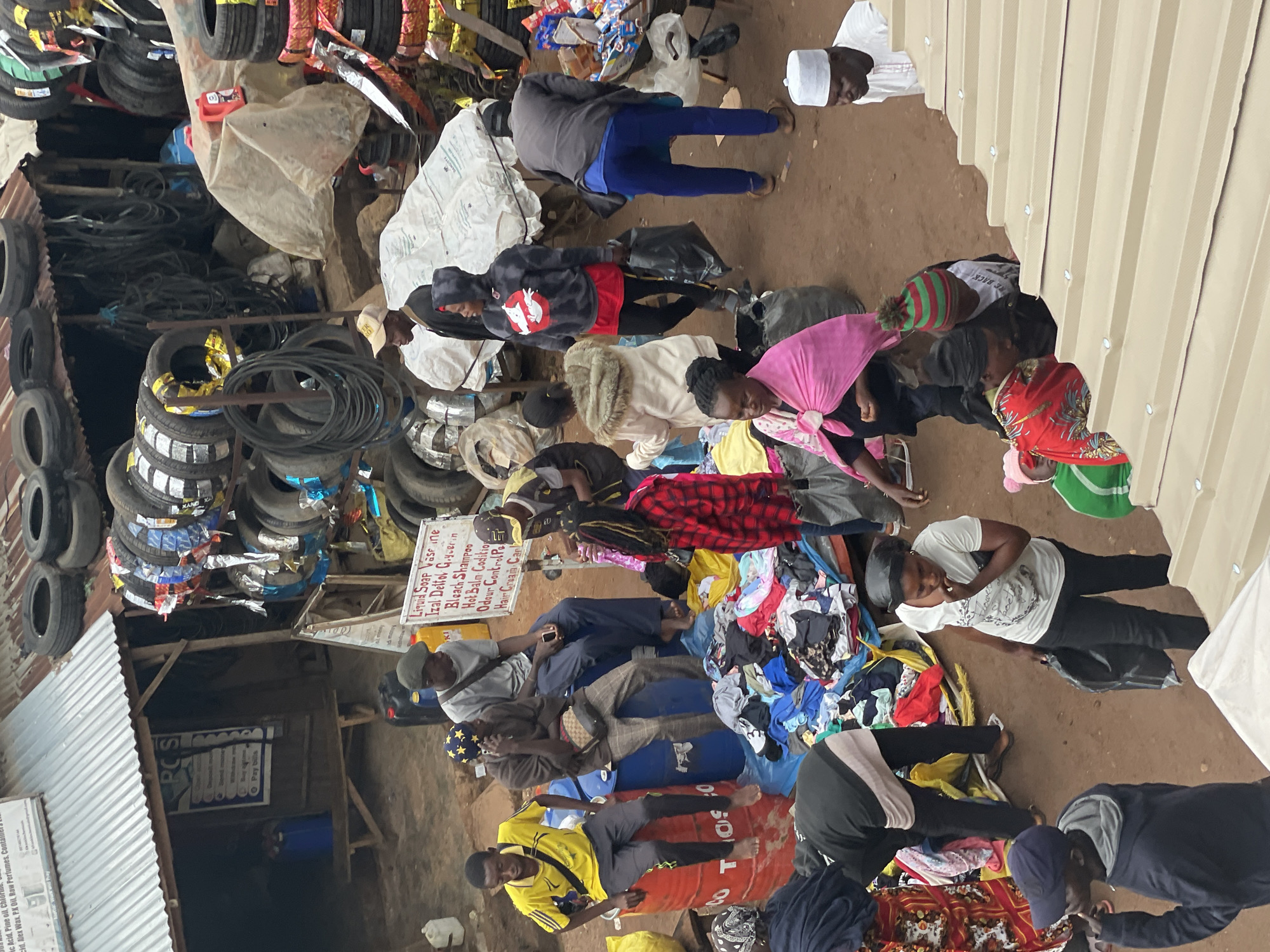 The image shows an outdoor market scene with a variety of goods, including tires stacked high and various clothing items spread on tarps. Several people are present, some sitting and others standing, engaged in activities suggestive of a marketplace.
In the foreground, a man wearing a dark blue hooded jacket and a baseball cap is looking to the left, with his left hand raised. To his right, a woman in a white t-shirt with a graphic print and black pants is holding a black plastic bag and looking down. Further right, a person wearing a pink headscarf and a light-colored jacket is visible. A child in a green and red striped hat is seated next to them.
In the middle ground, several individuals are interacting. One man in a yellow t-shirt and black pants sits on a red barrel. Next to him, a man in a brown jacket and plaid pants is on a blue barrel, talking on a phone. Another man in a gray t-shirt and dark pants is seated on a blue barrel, looking at a phone. A woman in a black hoodie with a red graphic and red pants walks with a black bag. A woman in a white coat with fur trim is standing. A woman in a red and black plaid jacket stands near a pile of colorful clothing.
In the background, more people are visible, and a sign with "PCS" is displayed on a building. There are numerous tires of various sizes stacked on racks and on the ground, indicating a tire vendor or related business. The ground is dusty earth. Some items are covered with tarps, and there are plastic containers and bags scattered throughout the scene. The overall environment suggests a busy, possibly informal, marketplace.