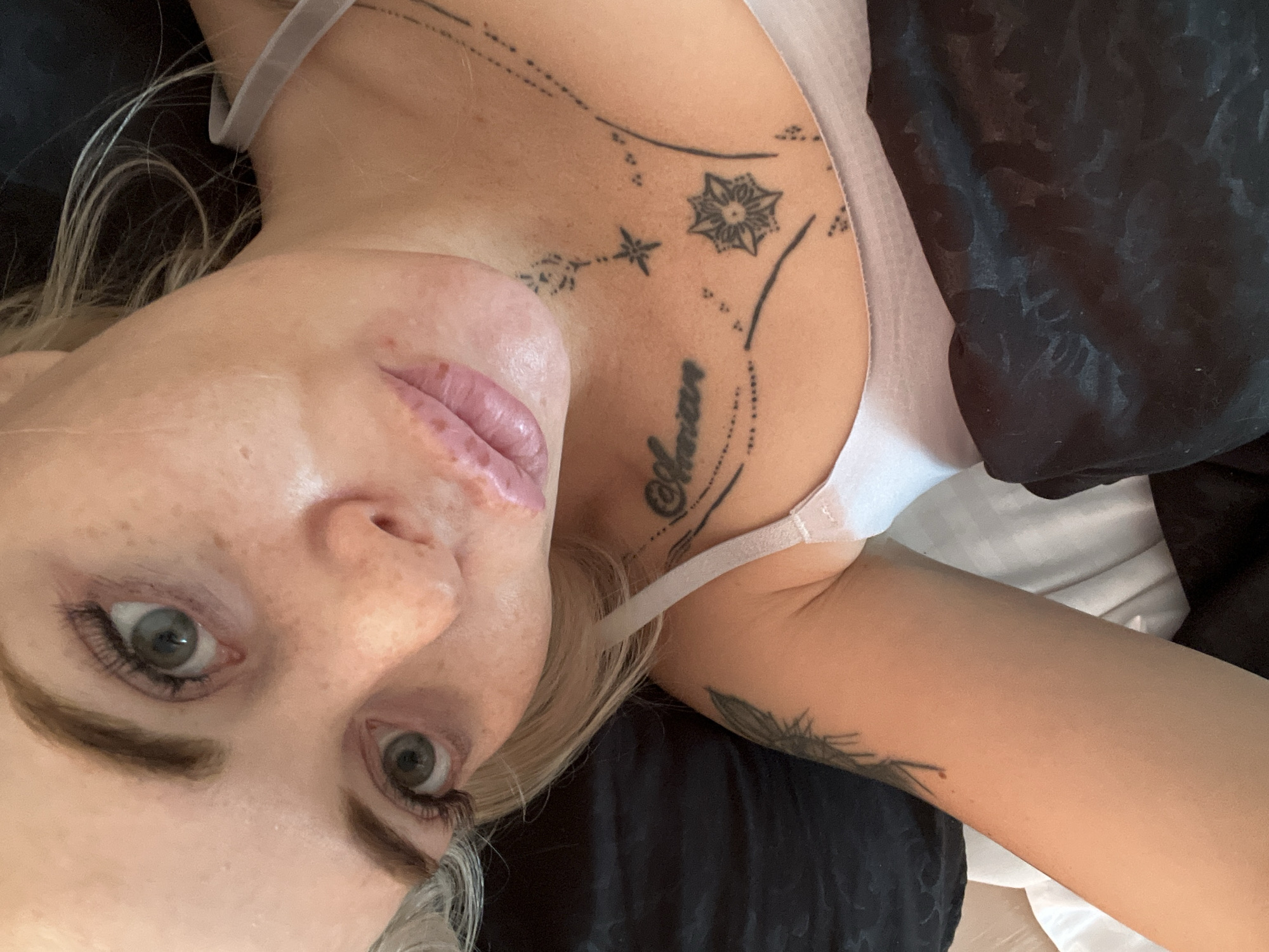 A close-up, overhead shot shows a person with blonde hair and light skin lying in bed. The person is wearing a light-colored bra and has several tattoos on their neck and arm. One tattoo on the neck appears to be text. The background consists of dark bedding and a white pillow. The lighting is soft and natural. The subject's eyes are open and looking towards the camera. The overall scene is intimate and relaxed.