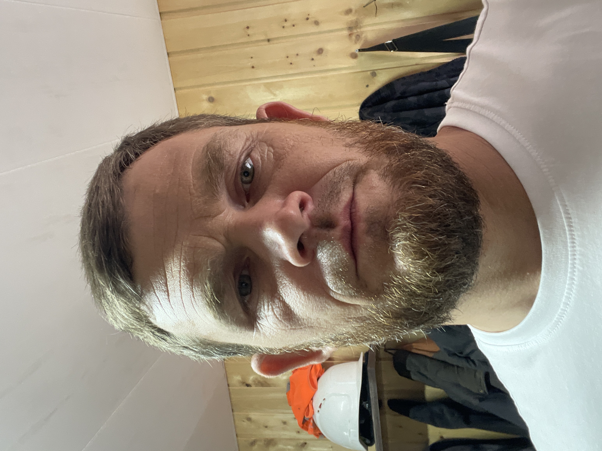 The media file is a close-up, slightly distorted selfie of a man. He has a beard and short, light brown hair. His expression is neutral, with his mouth slightly downturned. The man is wearing a white t-shirt. The background appears to be a room with wooden paneled walls. In the lower part of the image, there are some objects visible, including what looks like an orange safety vest and a white hard hat. The lighting suggests it is daytime. The scene appears to be indoors, possibly a workshop or storage area, given the presence of the safety gear. There is no visible text in the image. The overall impression is of a casual, informal self-portrait.