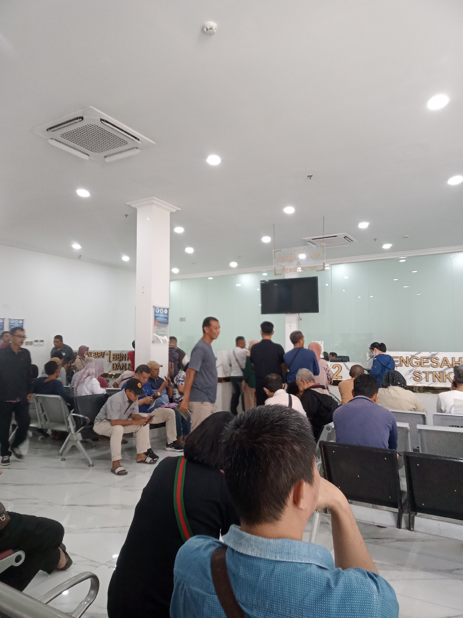 A wide-angle, eye-level shot captures a waiting area filled with people, likely at a government office or public service center in the city of Medan, Indonesia. The interior is brightly lit with fluorescent ceiling lights, and a ducted air conditioning unit is visible on the white ceiling. The walls are white, and a large glass partition separates the waiting area from a service counter. A flat-screen television is mounted on the wall behind the partition.
In the foreground, a person in a light blue collared shirt is seated, looking to the right with their hand resting on their chin. Behind them, another person wearing a black top with a green and red striped strap is seated, facing away from the camera.
The waiting area is furnished with rows of black chairs. Several individuals are seated, some looking at their phones, others engaged in conversation or simply waiting. People are dressed casually in a variety of clothing, including t-shirts, shirts, and headscarves. The floor is tiled with a light-colored marble pattern.
A sign above the service counter reads "LONET PENDAFTARAN & PENERIMAAN" which translates to "Registration & Reception Ticket." Another sign visible behind the glass partition says "PENGESAHAN STNK," indicating services related to vehicle registration renewal.
The overall impression is one of an organized but busy public space where people are waiting for their turn to be served. The presence of the "STNK" sign suggests this could be a Samsat office, a regional vehicle registration center in Indonesia.