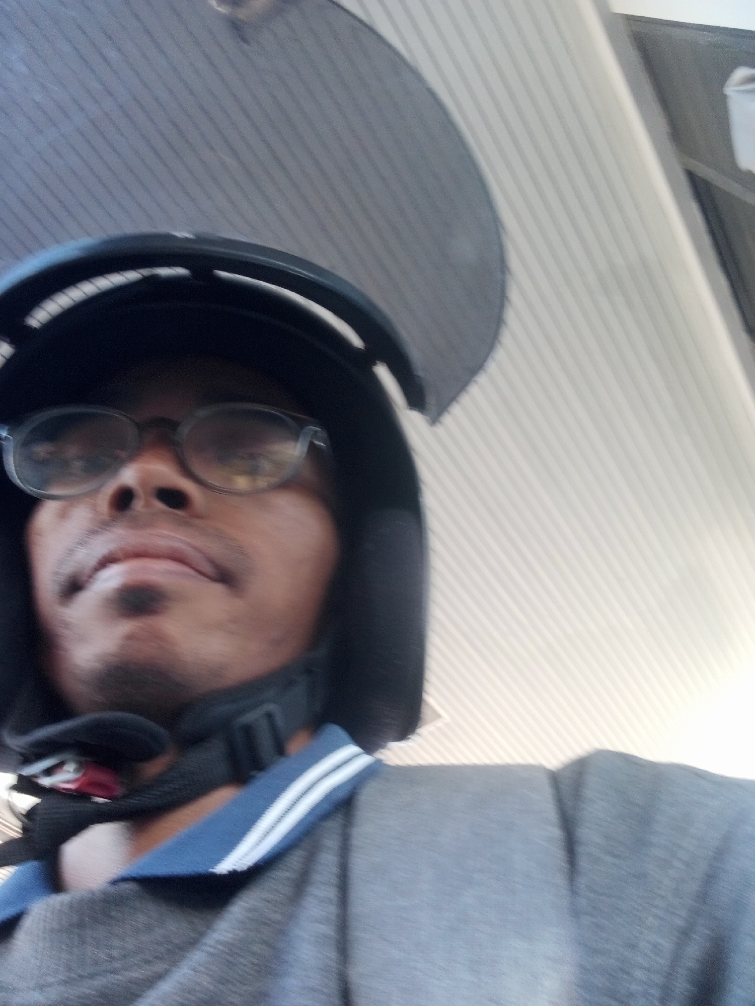 The image is a selfie taken from a low angle, showing a person wearing a black helmet and glasses. The person's face is partially obscured by the helmet. They are wearing a gray shirt with blue and white trim on the collar. The background appears to be an indoor space with a ceiling that has a striped pattern. There is no indication of the specific location in Sawotratap, Indonesia. The image is blurry and lacks clear details of the surroundings or any other individuals.