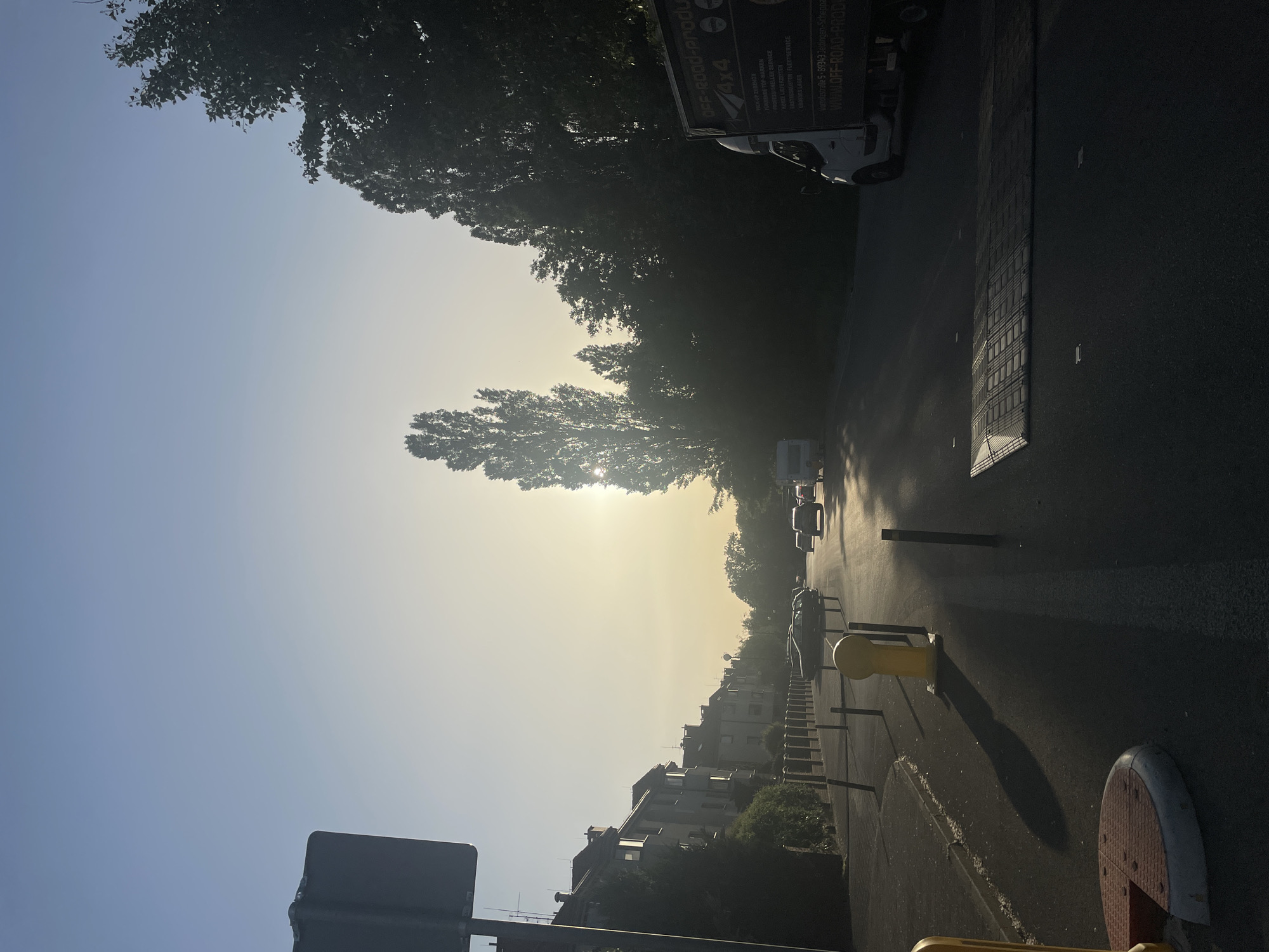 The image shows a street in an urban environment, likely taken in the early morning due to the low angle of the sun and the hazy quality of the light. The street is lined with trees on one side and buildings on the other. There are vehicles parked along the side of the road, including a truck with "OFF-Road-produ" printed on it. The text includes the phrase "Verholzstraße 5-89343 Jettingen-Scheppa," which is a location in Germany, not Wrocław, Poland. The scene also includes street furniture, like bollards, and road markings. The overall impression is of a quiet street at the beginning of the day.