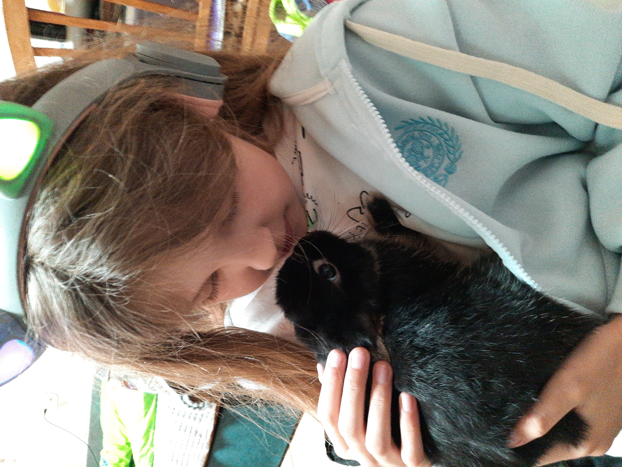 A child is cradling a black rabbit, seemingly interacting face-to-face. The child is wearing headphones with green accents, a light-colored hoodie, and a white top with a cartoon image, as well as a light-colored zipper hoodie. The child's hand holds the rabbit. The background is partially obscured, but what is visible indicates an indoor environment, including a wooden chair and other items. Location is not explicitly evident in the image and thus cannot be identified as Izhevsk, Russia.