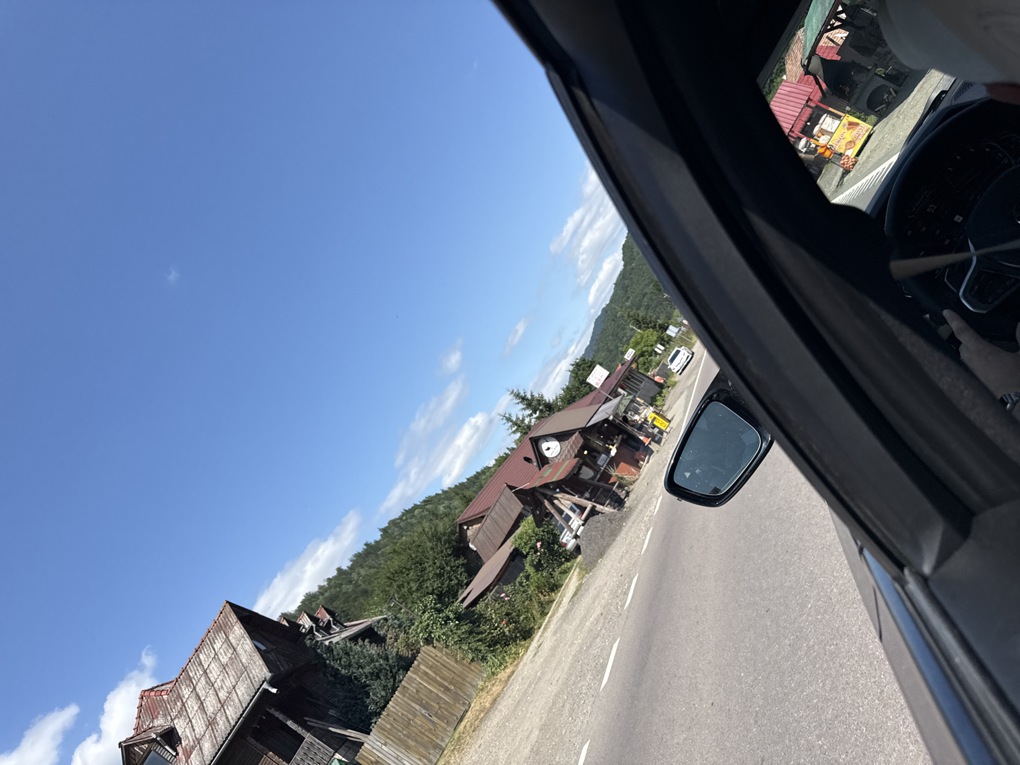 A street-level, angled exterior shot taken from inside a vehicle in Oituz, Romania, captures a scene under a clear blue sky with scattered clouds. The road in the foreground is asphalt with white lane markings. To the right, the interior of the vehicle is visible, including a dashboard display showing "53" and a steering wheel. The side mirror reflects a portion of the road and sky.
Adjacent to the road, on the left, is a traditional wooden house with a weathered red roof, situated on a hill surrounded by green trees. A wooden fence runs along the property. Further along the road, on the right side, a cluster of small, rustic buildings with red roofs line the street. One building appears to be a roadside food stall or restaurant, with visible signage and outdoor seating. There are a few vehicles parked or driving on the road, including a white car further down the street. The overall impression is a rural or semi-rural setting with a mix of natural landscape and small commercial establishments.