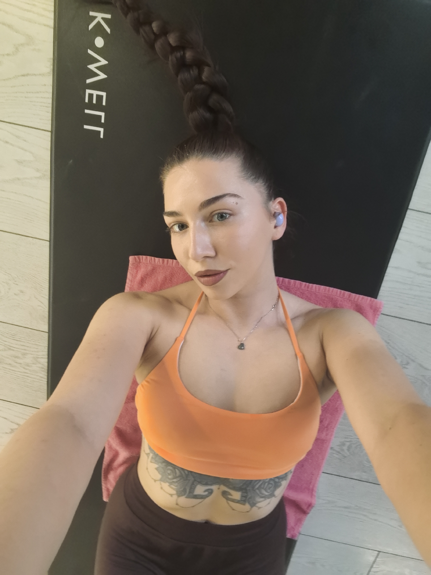 A woman is lying on a black yoga mat with the word "K-METT" in white text. She is wearing an orange sports bra and brown leggings. A pink towel is underneath her. She has a long braided ponytail and is wearing a blue earbud in her left ear. There is a floral tattoo on her abdomen, just below her navel. The floor appears to be made of light-colored wood planks.
The location is not identifiable as Stara Zagora, Bulgaria.