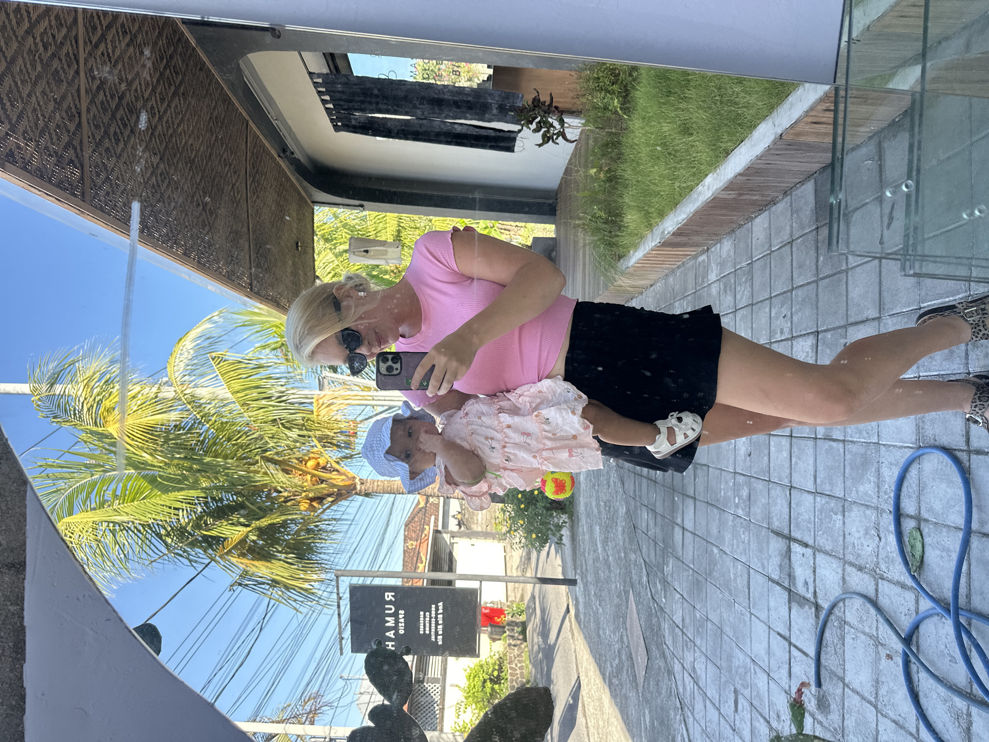 The image shows a reflection of a woman holding a baby in front of a glass surface. The woman is fair-skinned and has blonde hair. She wears sunglasses, a pink shirt, and a black skirt. She is holding a phone to take a picture of herself and the baby in the reflection. The baby is wearing a light pink dress with a cherry pattern and a light blue bonnet. Behind them, visible in the reflection, is a street scene, a palm tree, and a sign that says "RUMAH." The location is identified as North Kuta, Indonesia. There is paving stone on the ground on the right side of the image, as well as the presence of a blue hose.