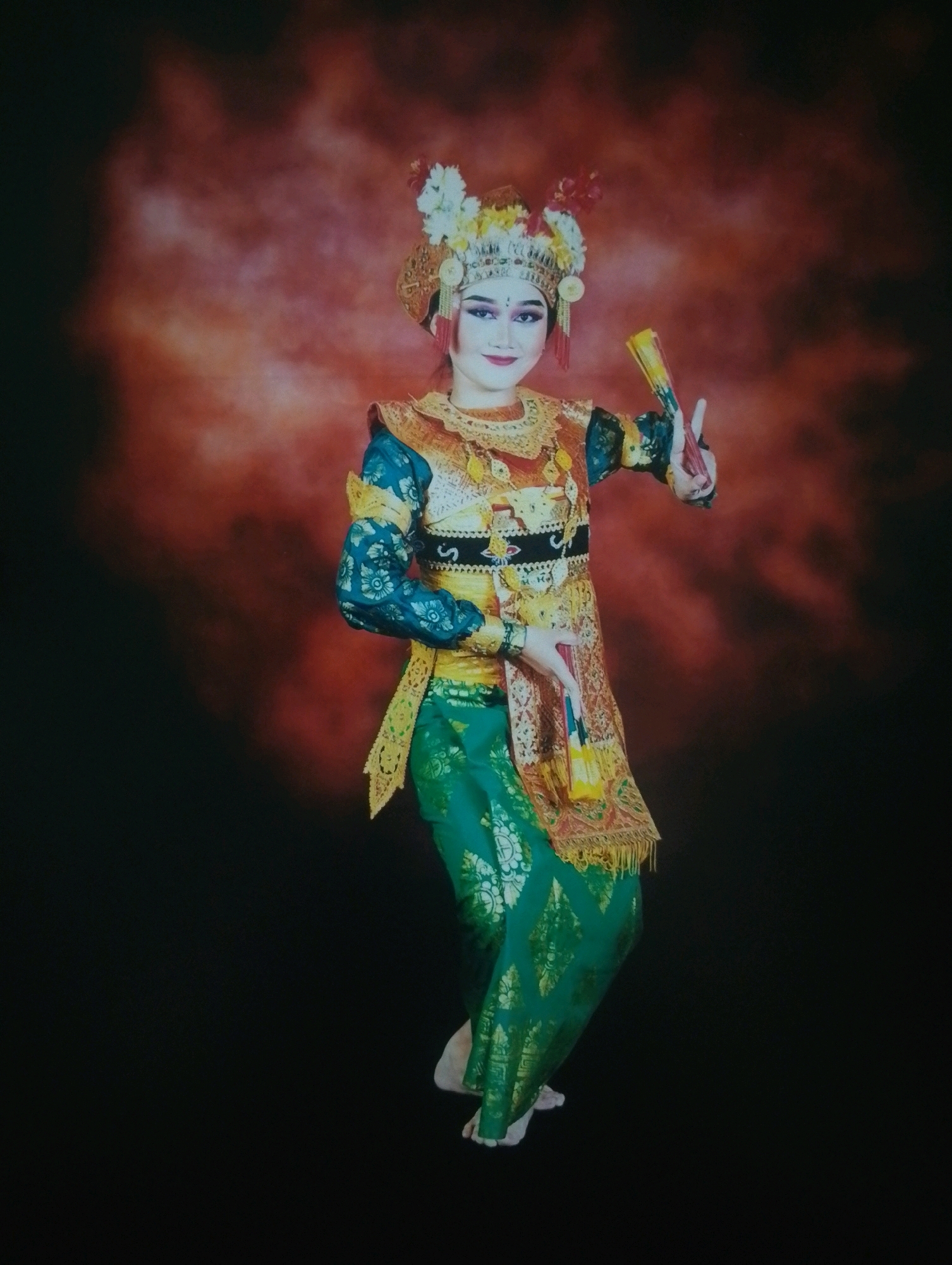 A full-body studio portrait shows a young Indonesian woman in traditional Balinese attire, performing a dance. She wears an elaborate headdress adorned with white and yellow flowers and golden ornamentation. Her costume includes a teal, long-sleeved top with floral patterns, a gold and red patterned sash, and a vibrant green sarong with gold motifs. The woman holds a colorful fan in her right hand and has her left hand raised. She has dramatic makeup, with dark eyeliner and red lipstick. The background is a gradient of dark brown at the bottom transitioning to a reddish-orange, mottled texture at the top. The lighting is focused on the subject, with the left side of the image darker than the right. This image was taken in the city of Depok, Indonesia.