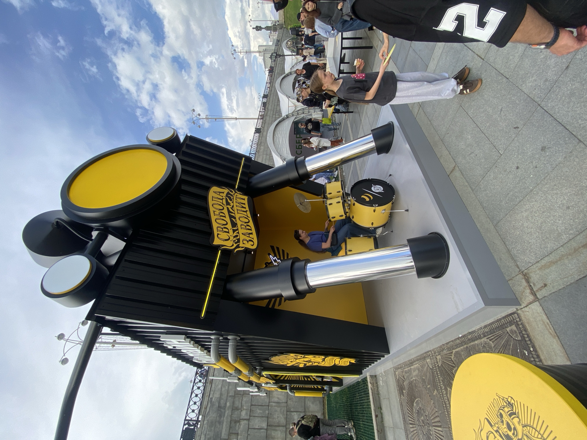 An outdoor scene features a large, interactive art installation resembling a giant drum kit, prominently displayed in black and yellow colors. The structure has large circular yellow elements at the top and two prominent silver cylindrical components extending outwards, mimicking oversized drum components. Inside this structure, a person wearing a blue t-shirt and jeans is seated, actively playing a smaller, real yellow drum set.
A young girl, wearing a black t-shirt and grey sweatpants, stands outside the installation, holding a yellow card or piece of paper and looking towards the drums. In the background, several other people are visible, some walking past and others gathered near white canopy tents, suggesting a public event or festival. The setting appears to be a city promenade or public square, with paved ground, street lamps, and a railing suggesting a waterfront or embankment in the distance. The sky above is bright blue with scattered white clouds, indicating good daytime weather.
Visible text on the main structure reads "СВОБОДА ЗАВОДИТ" (SVOBODA ZAVODIT), which translates to "FREEDOM STARTS/TURNS ON" or "FREEDOM IGNITES." A logo featuring a stylized skull with a crown is visible on a yellow circular element in the foreground. Another logo is present on the drum inside the installation. The overall atmosphere conveys curiosity and engagement with the unique musical display.