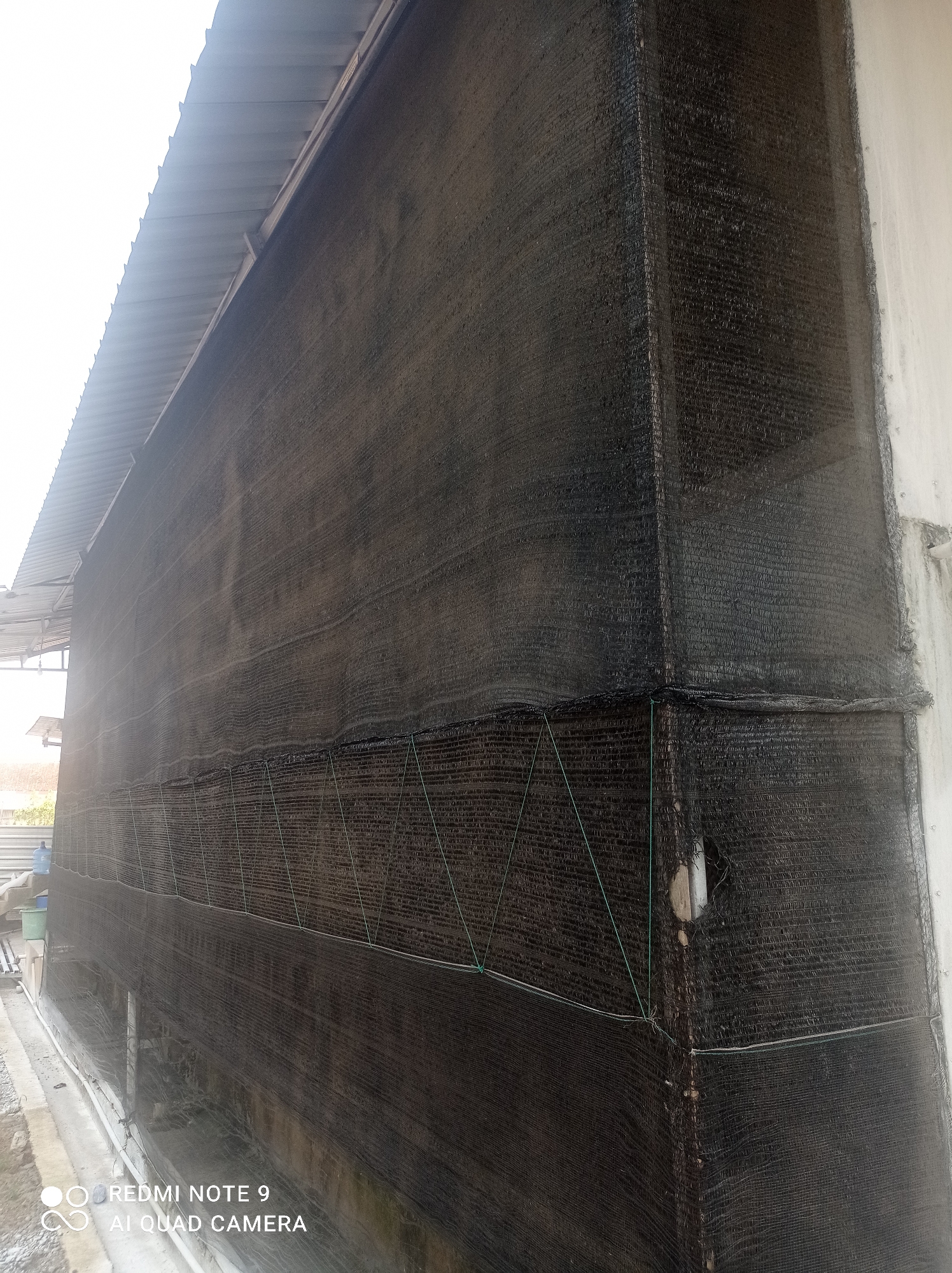 This image depicts the exterior of a building covered in black shade netting, located in Penaruban, Indonesia. The netting is attached to the building's walls and extends under a metal roof. The scene shows the corner of the structure, with one wall covered in netting that appears to be secured with green twine. The netting has some visible wear and tear, including a noticeable hole on the right side wall. The environment appears to be industrial or agricultural, with hints of outdoor elements like a clear sky and some vegetation in the background. There are no people visible in the immediate frame of the image.
