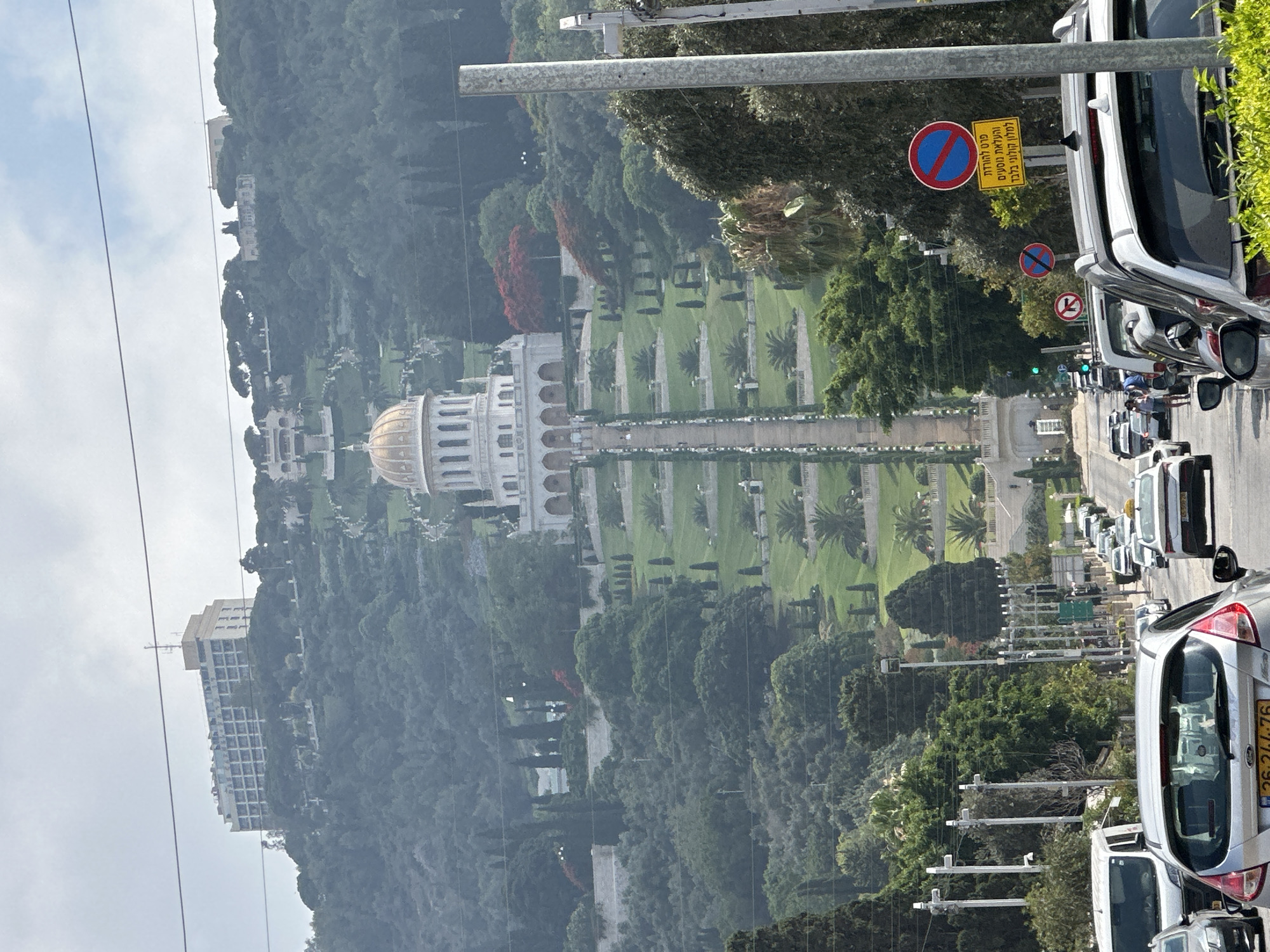 The image captures a view looking uphill towards the Baháʼí World Centre in Haifa, Israel. In the foreground, a traffic jam of cars is visible on a street, with a silver Ford Fiesta in the immediate foreground. Several other vehicles, including white vans and taxis, are also present. Traffic signs, including "no parking" and "no stopping" signs in Hebrew, are mounted on poles along the roadside. Beyond the street, a tiered garden with lush green lawns, cypress trees, and palm trees ascends the hillside. A grand white building with a golden dome, identified as the Shrine of the Báb, is prominently situated on the slope. Further up the hill, another large building, possibly a hotel, is visible. The sky is overcast.