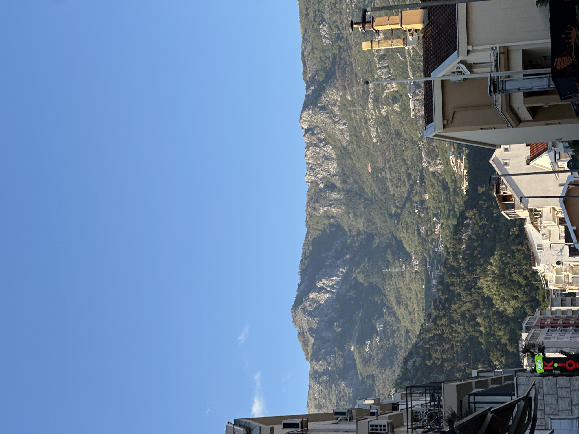 This image presents a panoramic view of Budva, Montenegro, showcasing the town's urban landscape set against a dramatic mountain backdrop.
In the foreground, at the bottom of the frame, stand several multi-story buildings. They feature light-colored facades and reddish-brown tiled roofs, typical of Mediterranean architecture. Details such as balconies, windows, and external air conditioning units are visible on these structures. On the bottom right, a prominent building displays a bright green and red sign with the text "K-OUT," likely indicating a commercial establishment.
A rugged and expansive mountain range dominates the midground, stretching horizontally across the scene. Its imposing slopes are densely covered with vibrant green trees and vegetation, interspersed with significant patches of exposed grey rock formations.
The background is filled with a clear, brilliant blue sky, which occupies the upper portion of the image. A few small, wispy white clouds are scattered near the mountain peaks, adding texture to the otherwise cloudless sky.
The bright, even illumination and the clear skies suggest a sunny day, likely mid-morning or early afternoon. No specific activity or event is depicted, as the image is a static landscape and cityscape photograph. The scene captures the picturesque juxtaposition of coastal urban development with the natural grandeur of the Montenegrin mountains.