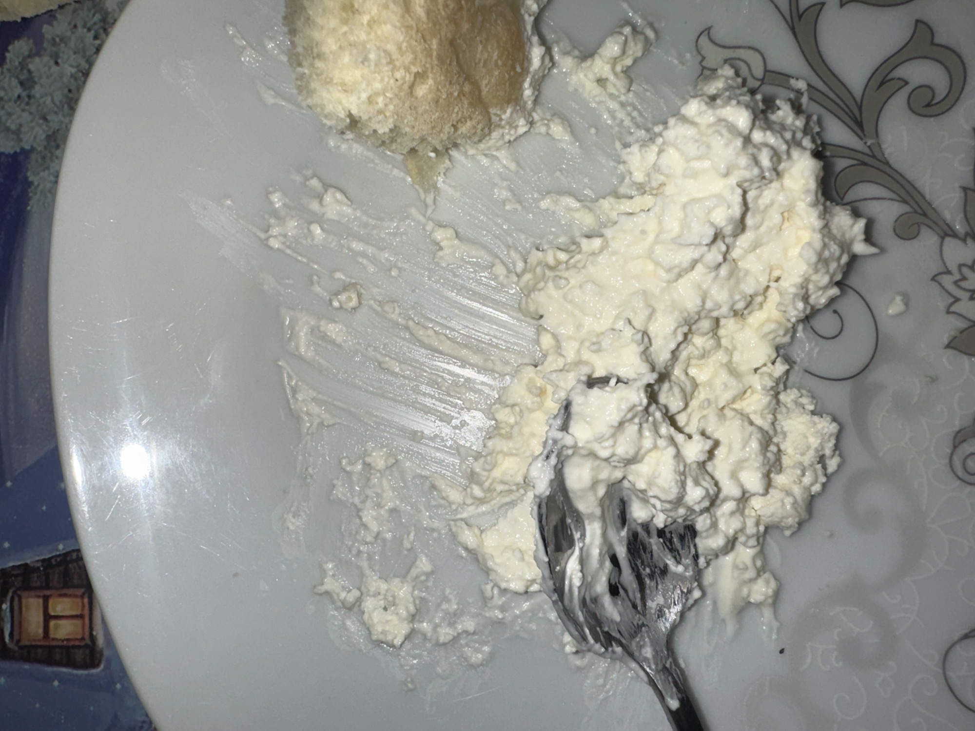A close-up, overhead shot shows a white plate with scattered dollops of cottage cheese and a fork. The plate is white with a subtle grey floral pattern around the rim. The cottage cheese is lumpy and moist. A fork, its tines visible, is partially submerged in a pile of cottage cheese, appearing to be in the process of scooping or mixing it. To the right, a small, golden-brown baked good, possibly a piece of bread or pastry, is also on the plate. The background of the image displays a tablecloth with a whimsical winter scene, featuring a small, glowing window in a dark building and stylized white trees against a blue sky. The lighting is bright, creating reflections on the surface of the plate.