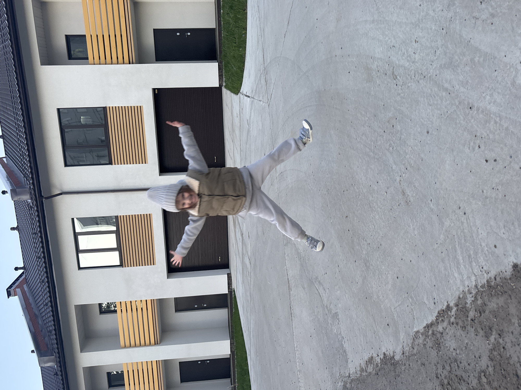 A young person wearing a hat, vest, long-sleeve shirt, and sweatpants is captured mid-air, appearing to be jumping. The background reveals modern residential buildings with distinct architectural features, including vertical wooden slats framing windows and balconies. The scene appears to be located in Chaikovsky, Russia. The ground consists of a concrete surface that is cracked in some places and covered with some debris.