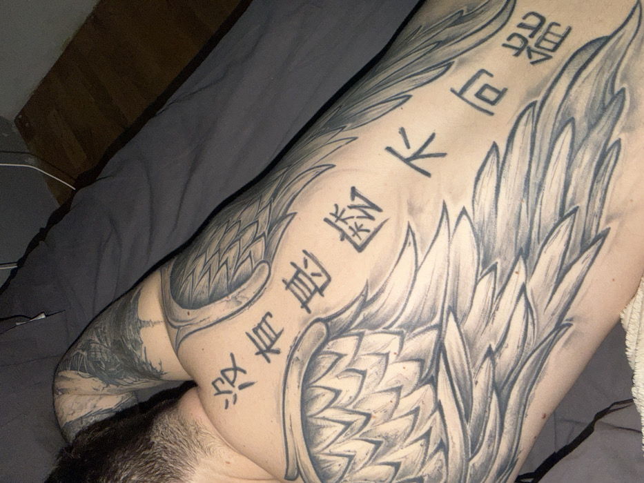 A close-up captures a person reclining on a gray bed, showcasing a shoulder tattoo featuring intricate wing designs intermingled with vertical lines of Asian script. The tattoo extends across the shoulder and upper back. The image is dimly lit, focusing on the tattoo's details and the individual's dark hair.