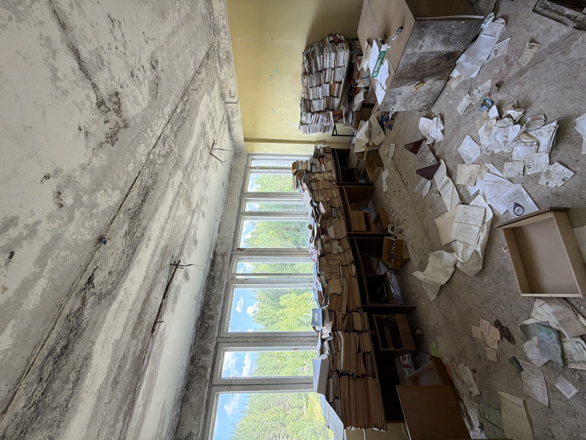 The image depicts an abandoned room, likely a former office or archive, within a building in Nikolsky, Russia. The room is filled with stacks of papers and documents, piled high on desks and on the floor. The papers appear aged and disheveled, with many scattered around the room. The overall environment suggests neglect and disuse, with peeling paint on the ceiling and walls, and debris on the floor. Through the large windows, a dense forest can be seen, indicating the building's location outside of a dense urban area. There are no people visible in the scene.