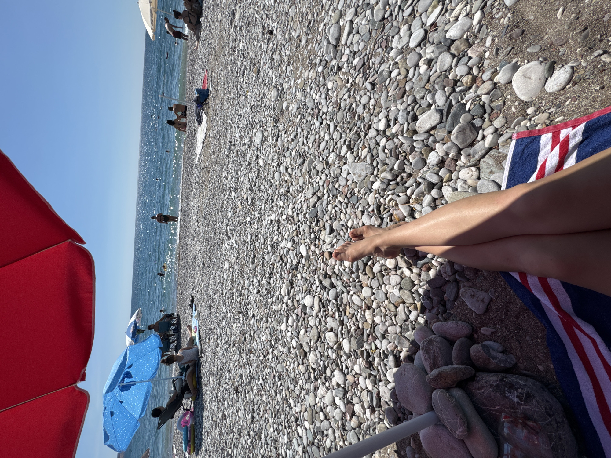 The image shows a pebble beach with people relaxing under umbrellas and swimming in the sea. In the foreground, a person's legs are visible, resting on a towel. The sea is calm and the sky is clear blue, indicating a sunny day. The location appears to be Šušanj, Montenegro, a coastal town known for its beaches. There are several umbrellas providing shade, and some beach chairs are set up on the shore. People are scattered across the beach, with some lying down and others wading in the water. The environment is a typical summer beach scene, with the sound of waves likely present.