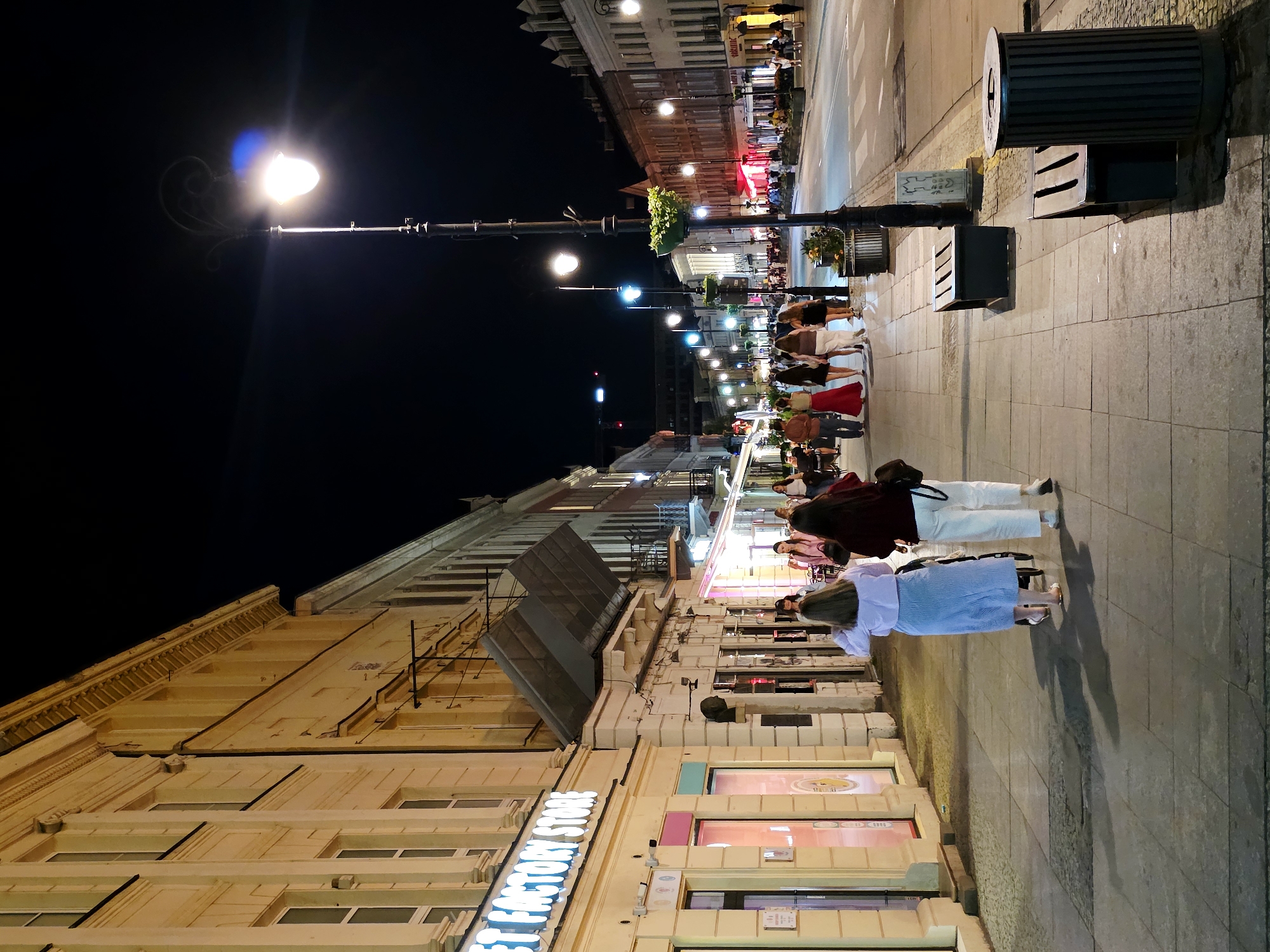 The image depicts a nighttime scene on a city street in Warsaw, Poland. The street is lined with buildings, some of which appear to be shops with illuminated signs, such as "SWEET FACTORY STORE." Several streetlights illuminate the pedestrian walkway and the road. A group of people are walking along the sidewalk, some in conversation and others with their backs to the camera. One individual is pushing a stroller. The atmosphere suggests a lively urban environment, possibly a commercial or entertainment district, with pedestrian activity continuing into the night.