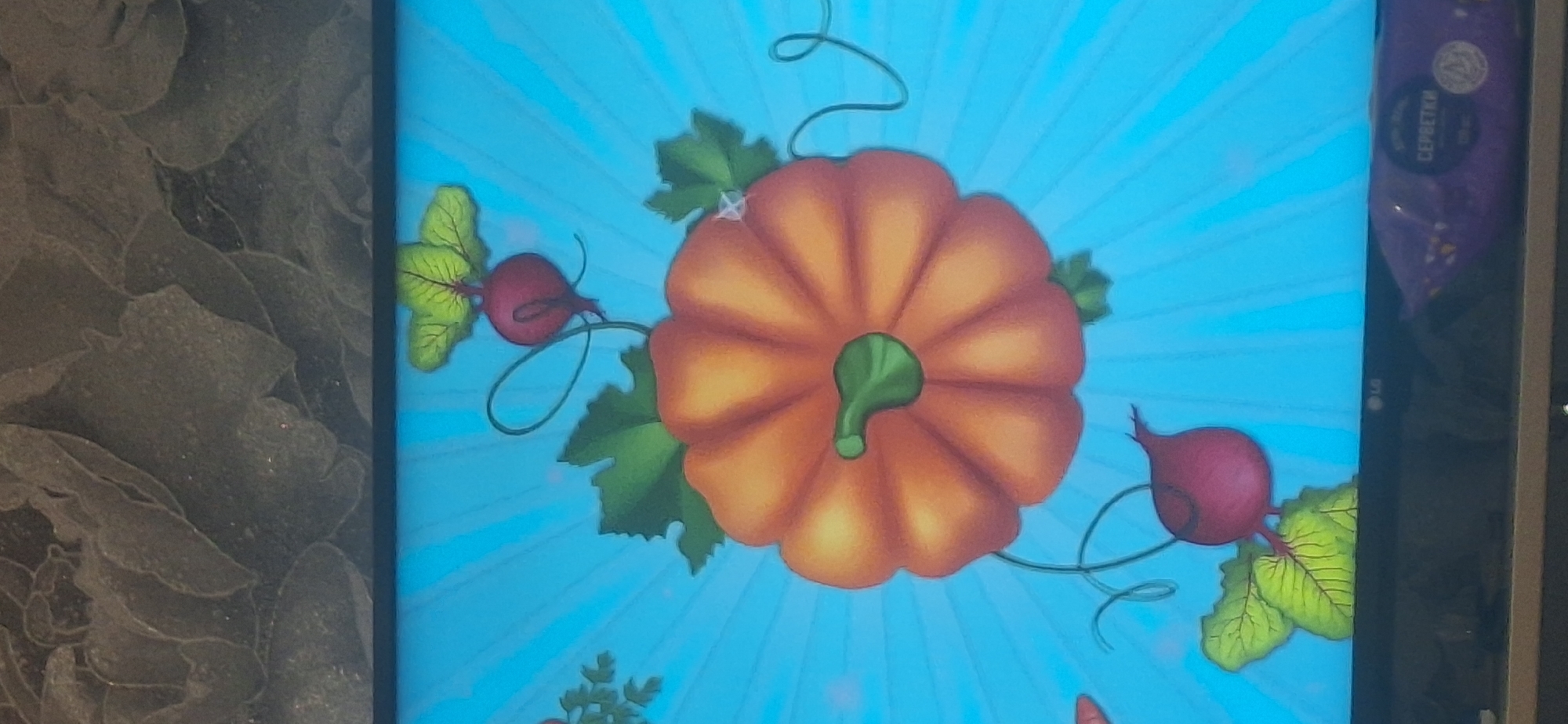 The image displays a digital rendering of vegetables against a bright blue background with radiating lines. A large, orange pumpkin is prominently featured in the center. To the upper right and lower left of the pumpkin are red beetroots with green leaves. There are also hints of other vegetables on the left side of the image, appearing as green leaves. The overall scene appears to be a still image from a screen or monitor. Below the screen, a dark surface is visible, on which a purple package with text in Cyrillic, including the word "СЕРБЕТКИ," is placed. The upper portion of the image shows a textured, dark gray surface, possibly wallpaper or a screen protector, with a floral pattern. The scene is static and does not contain any people or specific geographic markers indicating a location in Bila Tserkva, Ukraine.
