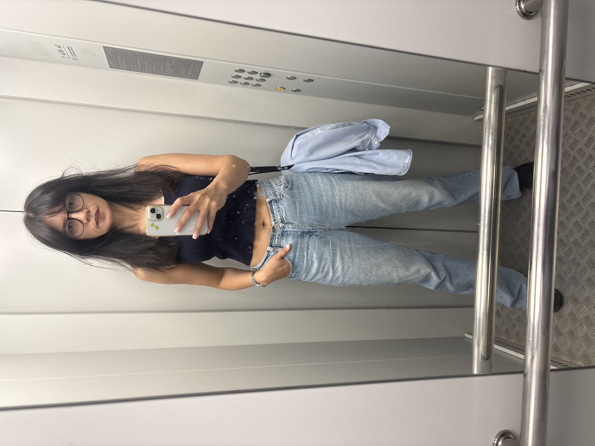 A woman with dark hair and glasses stands in an elevator, taking a mirror selfie with her phone. She is wearing a dark sleeveless top, light-wash jeans, and black shoes. A light blue shirt is draped over her left shoulder. The elevator has silver walls, a metal handrail, and a diamond plate floor. There are elevator buttons visible on the right wall. The image is a full-body shot, showing the woman from head to toe. The setting appears to be an indoor elevator, and the specific location in Chaikovsky, Russia cannot be determined from the visual information provided.