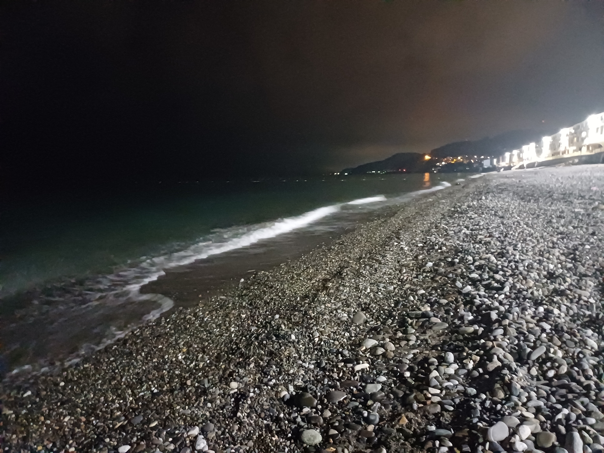 The image depicts a nighttime scene of a pebbled beach along a coastline in Sochi, Russia. In the foreground, a vast expanse of wet and dry dark gray, white, and black pebbles covers the beach, extending towards the viewer. The midground features the dark green-blue sea with foamy white waves gently breaking onto the shore, creating a luminous line across the frame. In the distant background, a dark mountainous landmass rises, illuminated by numerous blurry, warm-toned city lights strung along its base, indicating a coastal settlement. To the far right, a series of brightly lit, multi-story buildings, likely part of a resort or urban development, stand prominently along the coastline, reflecting light onto the water. The sky above is dark and cloudy, with a faint, diffuse orange glow visible near the horizon due to light pollution. No people or specific human activities are observable.