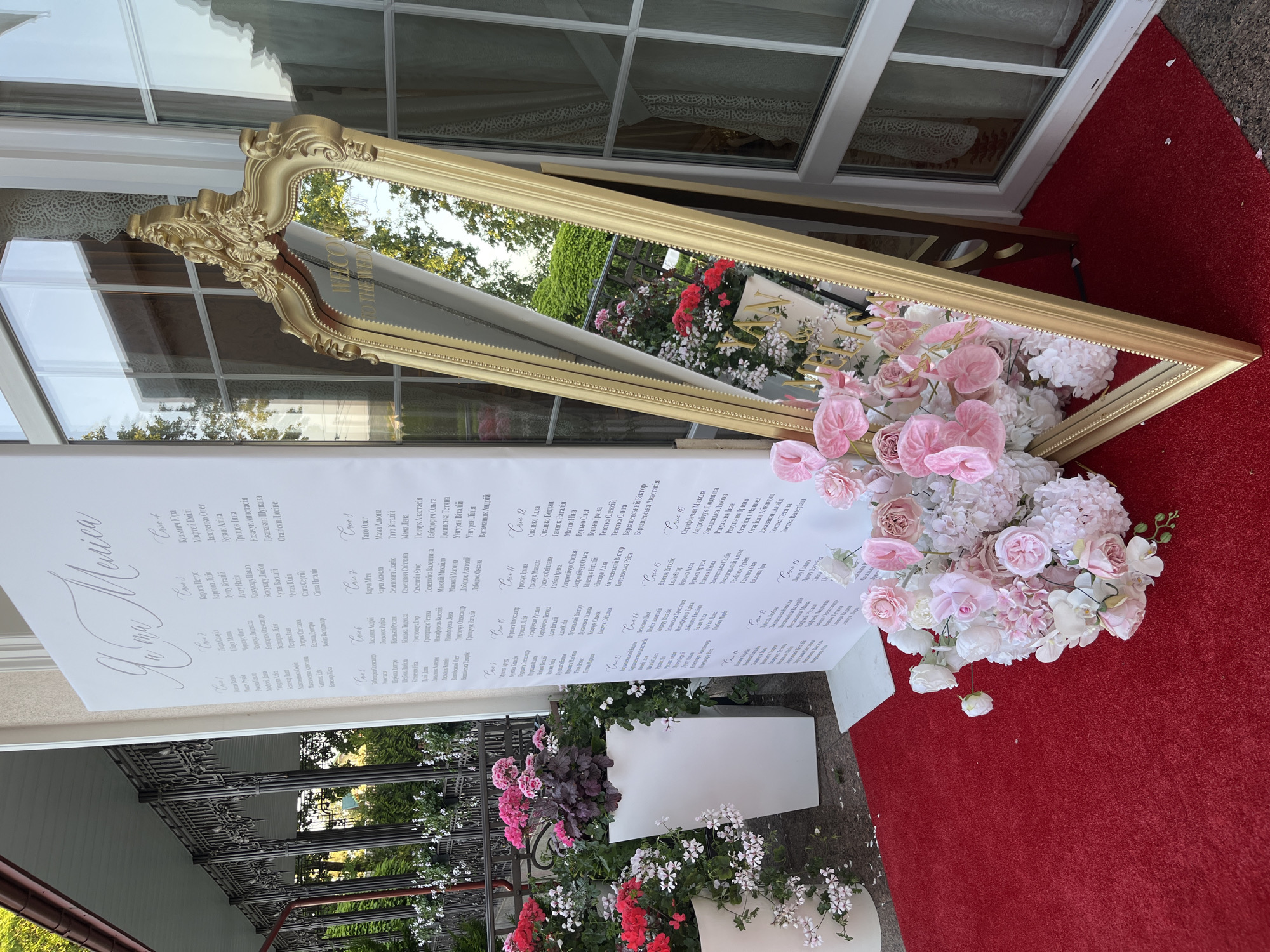 The image shows a wedding welcome sign and a mirror on a red carpet outside a building. The welcome sign is white with text in a script font, and appears to be a seating chart for a wedding. The mirror is ornate and gold, reflecting trees and the sky. There are also floral arrangements with pink and white flowers on the carpet. The location is identified as Dubivtsi, Ukraine.