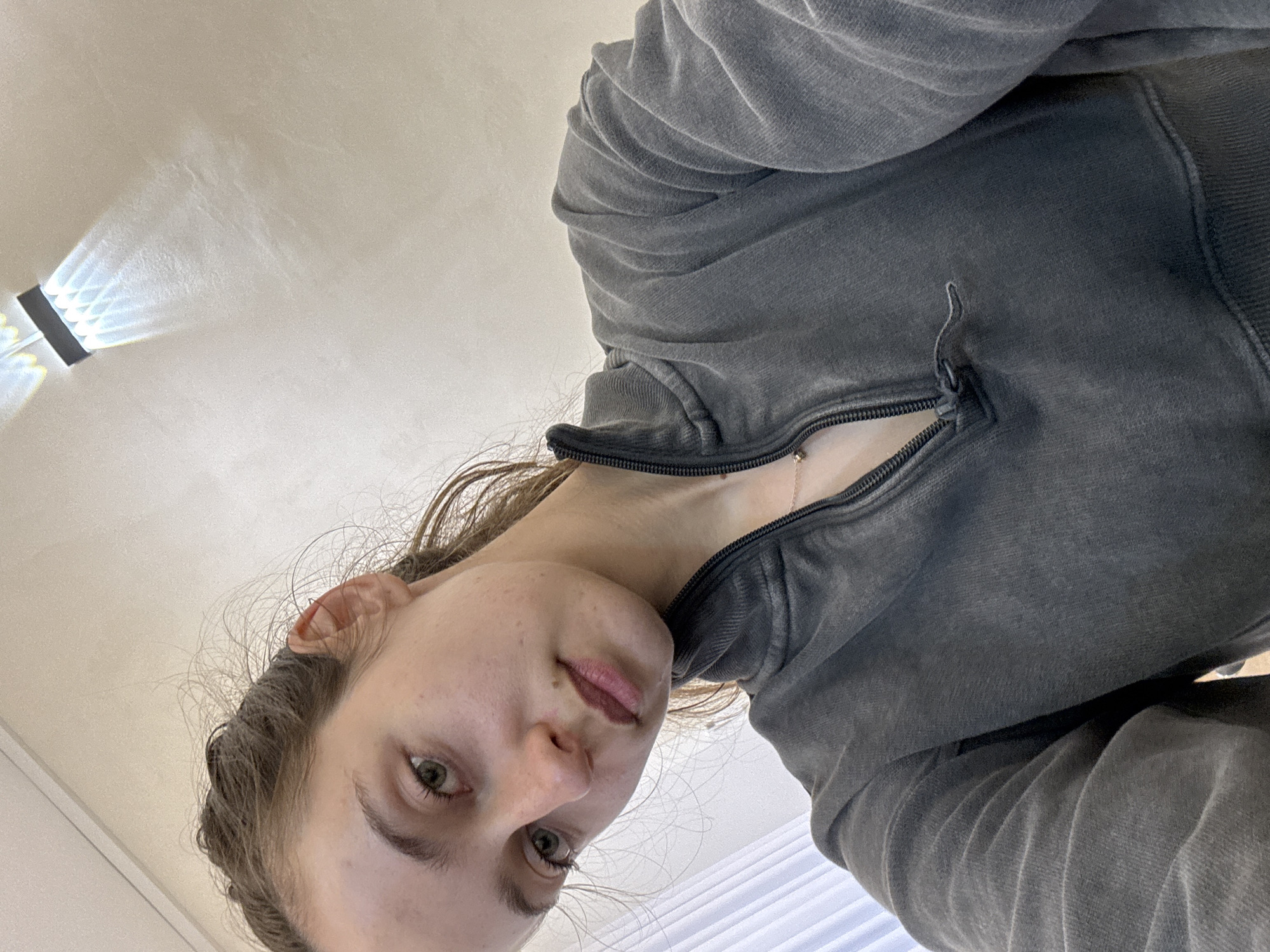 The image is a rotated, low-angle selfie of a young person, likely female, taken indoors. The subject is seen from a worm's-eye view, with the camera oriented horizontally, making the person appear on their left side.
The person has fair skin, light brown hair pulled back, and green or hazel eyes, looking directly into the camera. Their expression is neutral to slightly contemplative. They are wearing a dark grey or charcoal-colored hooded sweatshirt with a zipper partially undone, revealing a light-colored garment beneath and a delicate, silver-toned necklace with a small pendant. The sweatshirt fabric appears soft and possibly worn or faded.
The background consists of a light-colored ceiling. In the upper left, a light source or fixture creates distinct, angular patterns of bright light and reflections on the ceiling, possibly due to a prism or textured surface. In the lower right, white horizontal blinds or curtains are partially visible. The lighting is bright, suggesting daytime. No text is visible in the image. The depicted scene is an indoor environment, located in Ramat Gan, Israel.