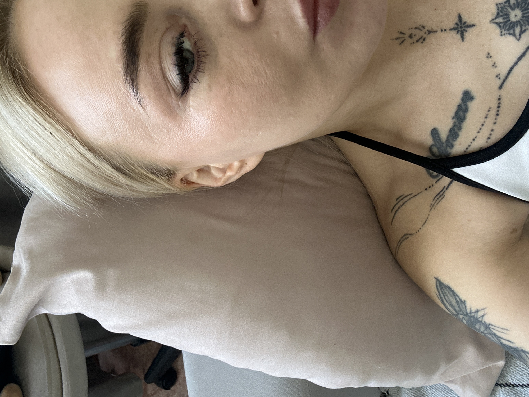 A close-up image shows a woman's face and upper body resting on a pillow. She has blonde hair, visible eye makeup, and several tattoos on her neck and upper chest. Tattoos are visible on her upper chest and arm. The background includes a pillow and a glimpse of a chair. The image does not depict any identifiable location.