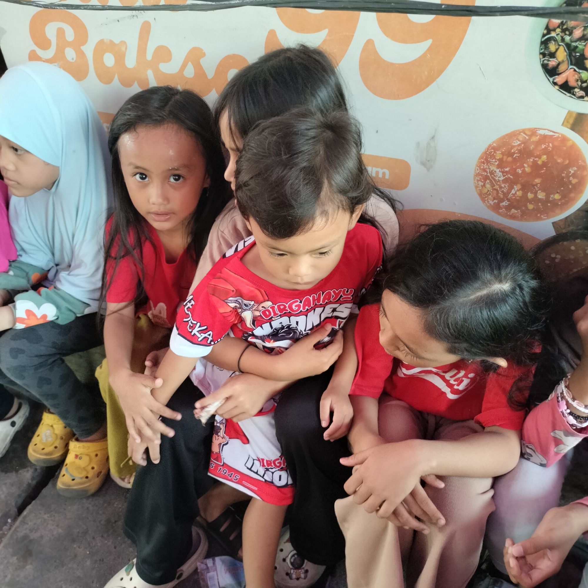 A group of young children are seated together. The child in the foreground wears a red t-shirt with graphics. To their left, another child is wearing a blue headscarf and light blue shirt. Other children in red t-shirts are also visible. Their hands are interlocked and resting on their laps. The background appears to be an outdoor setting, with a wall featuring orange and white lettering and images of food. The lettering suggests the location is related to "Bakso", a type of Indonesian meatball soup. Based on the distinct style of the "Bakso" signage and the overall ambiance, the scene is likely a street-side eatery or a food stall in Tangerang, Indonesia.