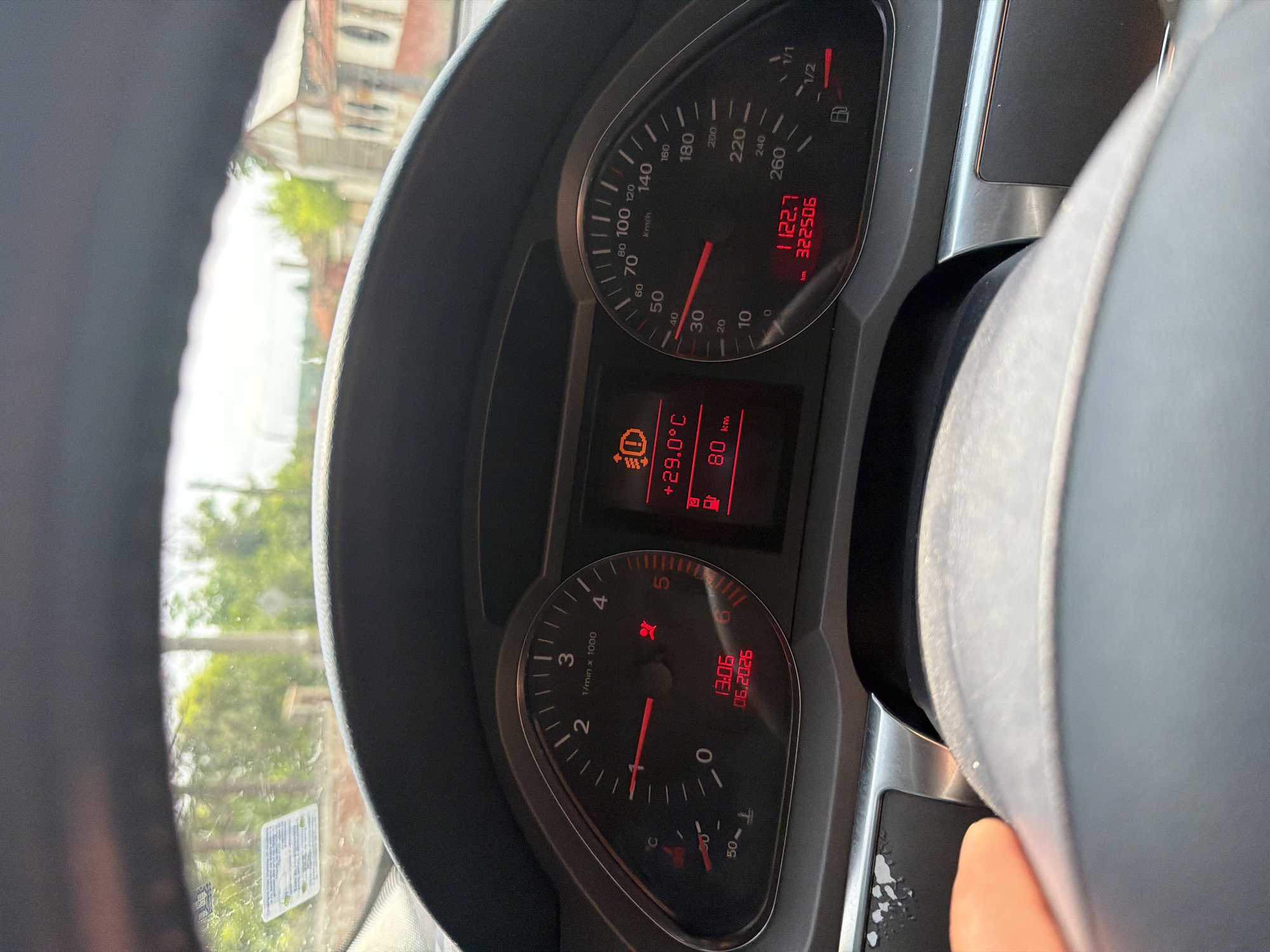The image presents a first-person perspective from the driver's seat of an Audi vehicle, specifically focusing on the dashboard and a partial view of the exterior through the windshield. A human hand is visible on the lower left portion of the steering wheel.
The dashboard instruments are illuminated in red against a dark background. The tachometer on the left indicates approximately 1000 RPM. Below it, a temperature gauge shows a low reading. Two red warning lights are active on the tachometer face: one for the airbag system and another for the seatbelt. A digital display within the tachometer reads "13:06" and "06.2026," likely indicating the current time and a date.
The central digital display provides further information: the ambient temperature is +29.0°C, and the remaining fuel range is 80 km. A yellow warning light, depicting an exclamation mark within a circle and brackets, is illuminated, signifying an issue with the brake system.
The speedometer on the right indicates a speed of approximately 30-35 km/h. Below the speedometer, a digital readout displays a trip mileage of "1122.7 km" and an odometer reading of "322506 km." The fuel gauge shows the tank to be approximately one-quarter full.
Through the windshield, a bright, sunny outdoor scene is visible. The immediate view comprises green foliage, trees, and a utility pole. In the mid-distance and background, several buildings with traditional reddish-tiled roofs and light-colored facades are discernible, consistent with the architectural style found in the city of Basarbovo, Bulgaria.