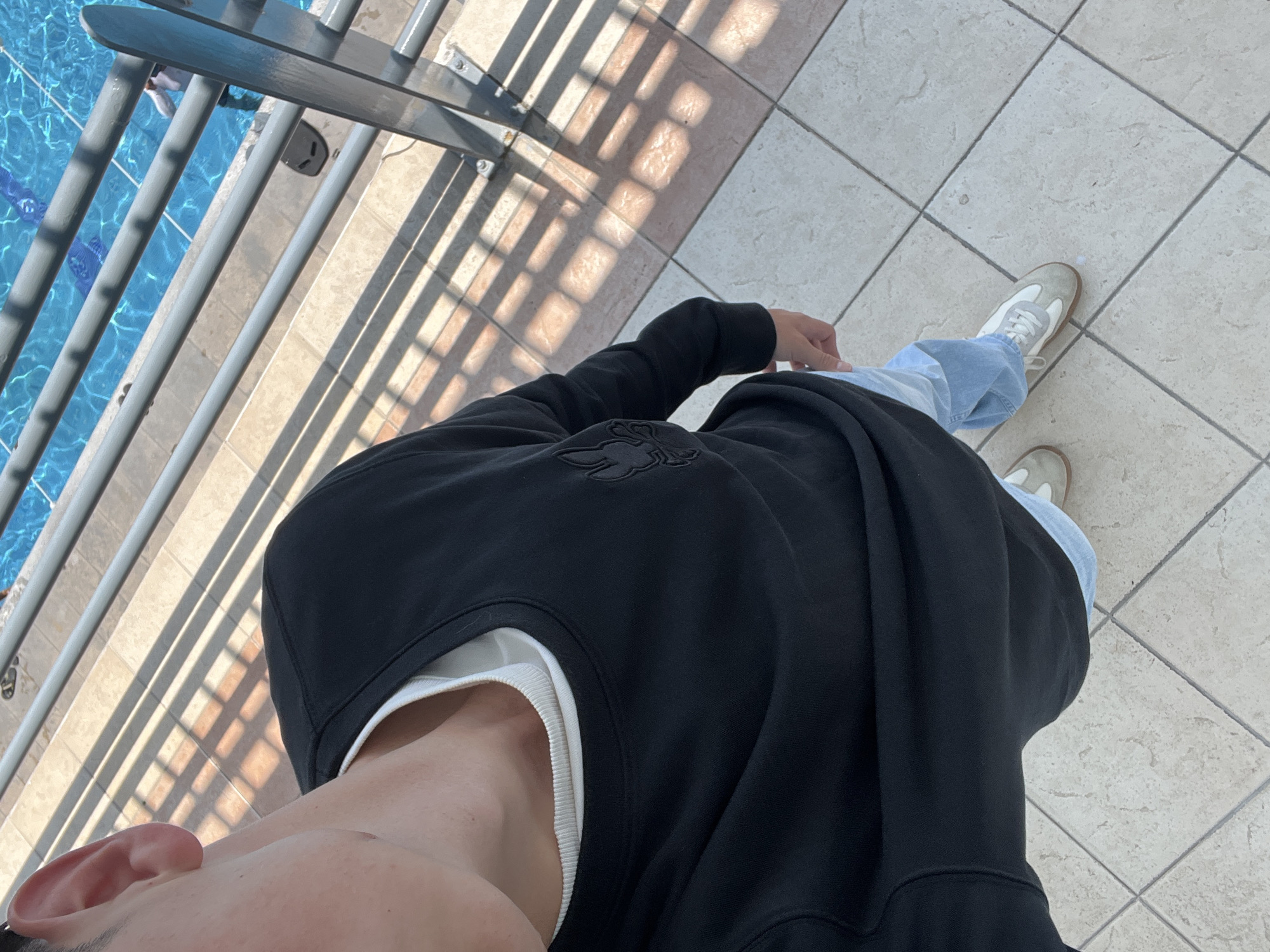 The image shows a person standing near a railing, likely by a pool. The person is wearing a black sweatshirt with a logo, a white undershirt, light blue jeans, and light-colored sneakers. The railing is metal and casts shadows on the tile floor. The pool is visible in the background. The location is Unknown, Unknown.