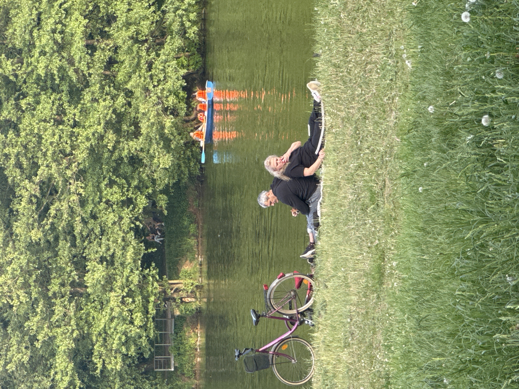 The image shows a scene by a river or canal. A dense green tree line runs along the left side of the image. On the river, a blue boat with orange life jackets sits on the water, which reflects the surrounding scenery. On the right bank, the ground is covered with green grass and dandelions.
An elderly couple is sitting on the grass near the water's edge. A bicycle is parked nearby. The man is crouching, while the woman is leaning back, appearing relaxed.
The location is most likely in Moscow, Russia, as inferred from the composition, and the presence of a river landscape possibly similar to the Moscow River or its canals. However, specific landmarks or architectural features definitively confirming the location are not discernible from the image.