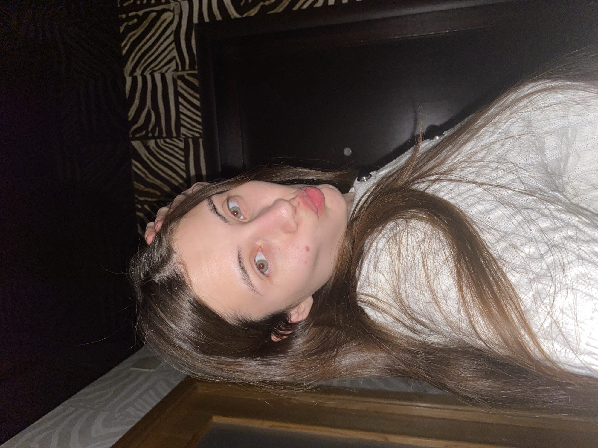 This media file captures a close-up, rotated portrait of a young woman in an indoor setting, likely a bedroom, within what is contextually identified as Votkinsk, Russia. The woman, appearing to be in her late teens or early twenties, has long, straight dark brown hair that cascades around her. She looks directly at the camera with wide, light green or hazel eyes and a playful expression, her lips slightly pursed in a "kiss face." A noticeable red blemish is visible on her left cheek. She is wearing a white, possibly knitted or textured, long-sleeved garment.
The background elements suggest a personal space. Directly behind her head is a dark, possibly wooden or upholstered, headboard. To the left, a wall is adorned with a distinctive black and white zebra-stripe pattern. A dark area, possibly the upper part of the wall or a dark piece of furniture, is visible above the patterned wall. At the bottom edge of the frame, a wooden surface or frame is present. The lighting in the image is dim, suggesting the photo was taken in low light conditions, possibly during the evening or night. There is no visible text in the image.