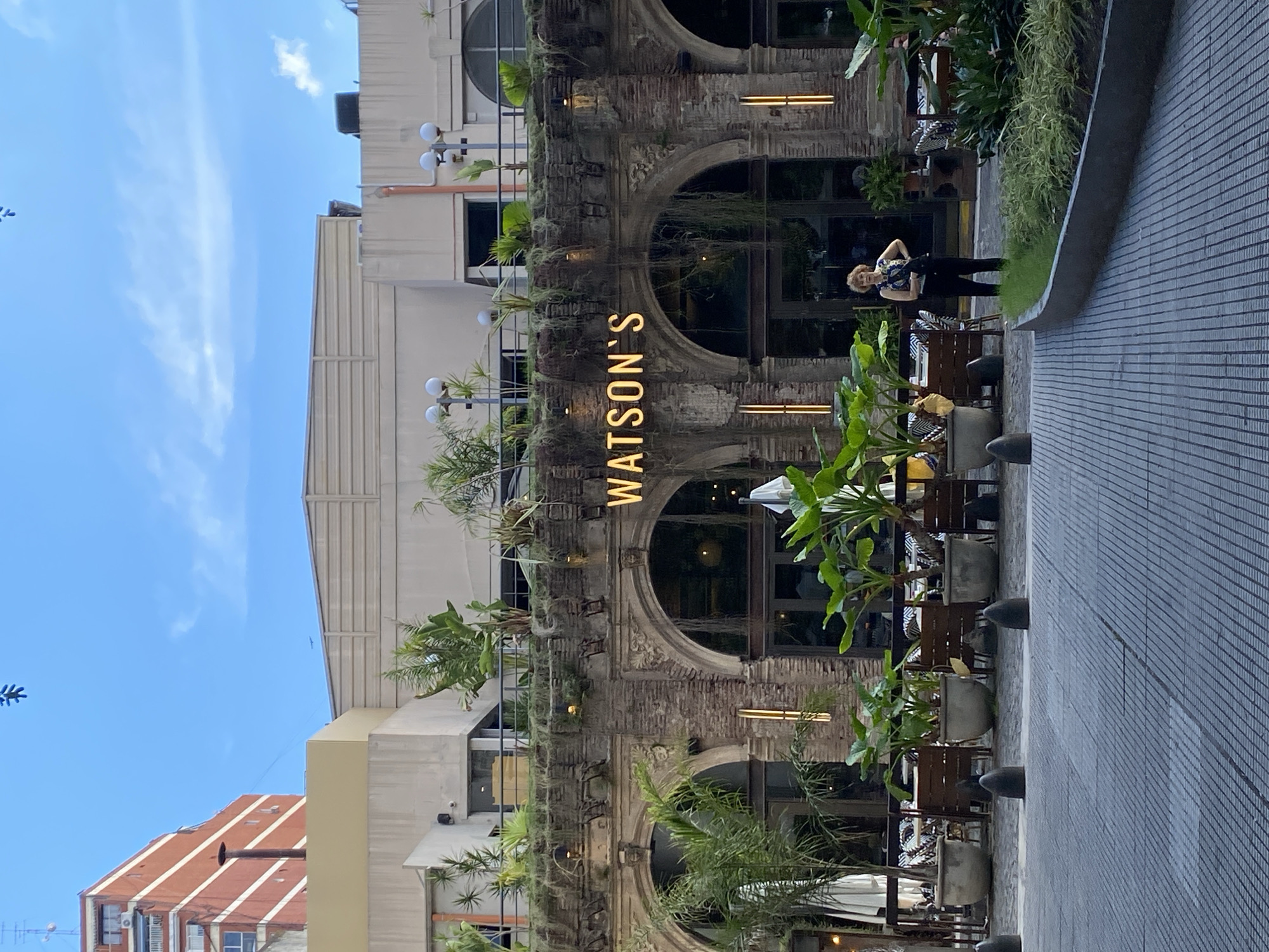 The image depicts the exterior of "Watson's" restaurant in Buenos Aires, Argentina. The restaurant's facade features a weathered brick structure with arched windows, adorned with various plants. A neon sign displaying the restaurant's name is vertically positioned on the facade. The outdoor seating area has wooden chairs and tables, interspersed with potted plants. A woman stands near the entrance. The sky is clear blue. The surrounding buildings are a mix of architectural styles. The ground is paved with gray tiles.