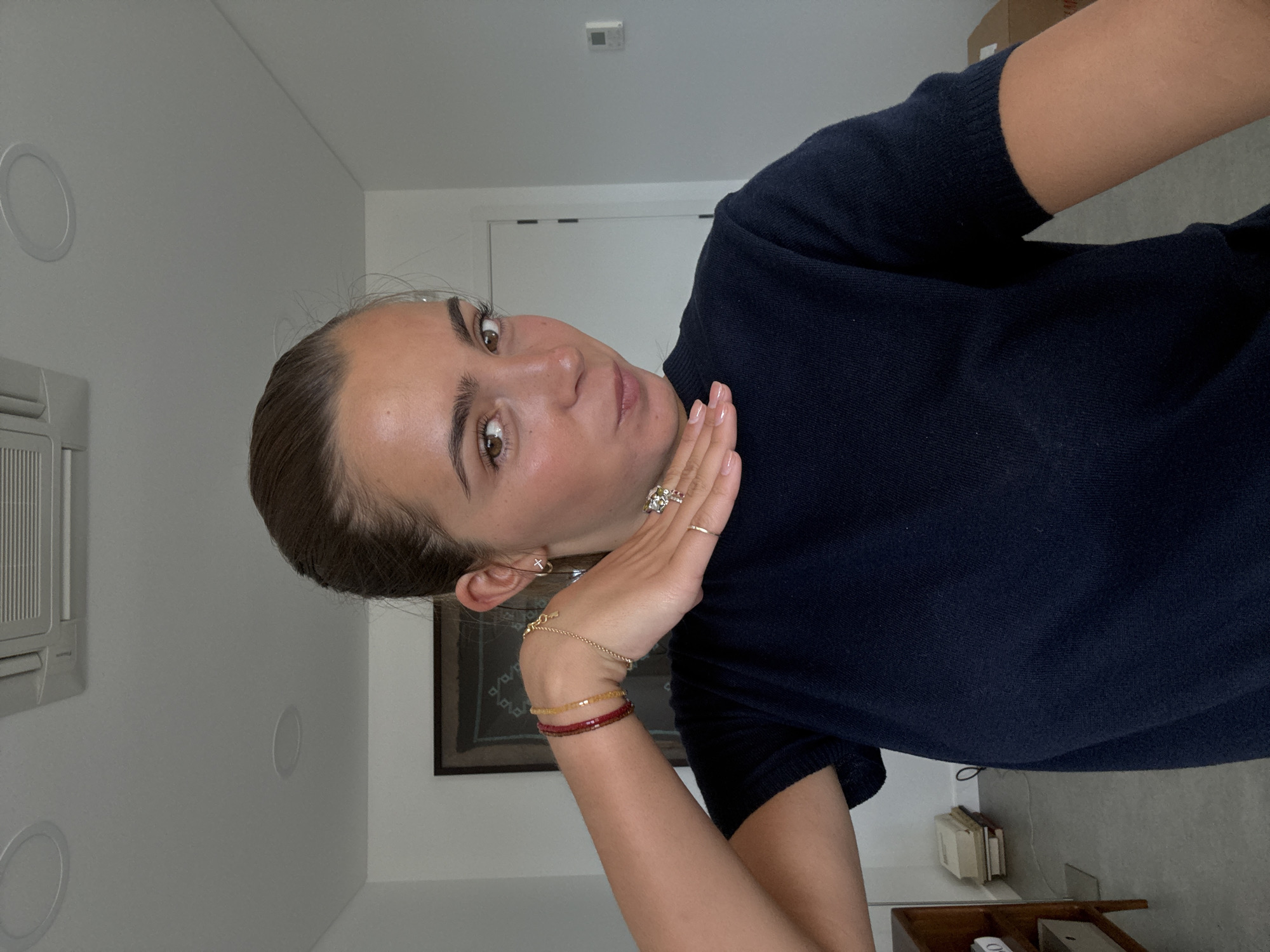 A woman poses indoors, looking at the camera with her right hand near her face. She wears a dark blue shirt, a gold bracelet, and multiple rings on her hand. Behind her is a white wall with a circular vent and a framed picture. A partially visible air conditioning unit is on the wall to her left. The room appears to be well-lit and tidy, with a doorway visible in the background. It is not possible to determine if the location is in Torres Vedras, Portugal, as the photograph does not provide the necessary context.